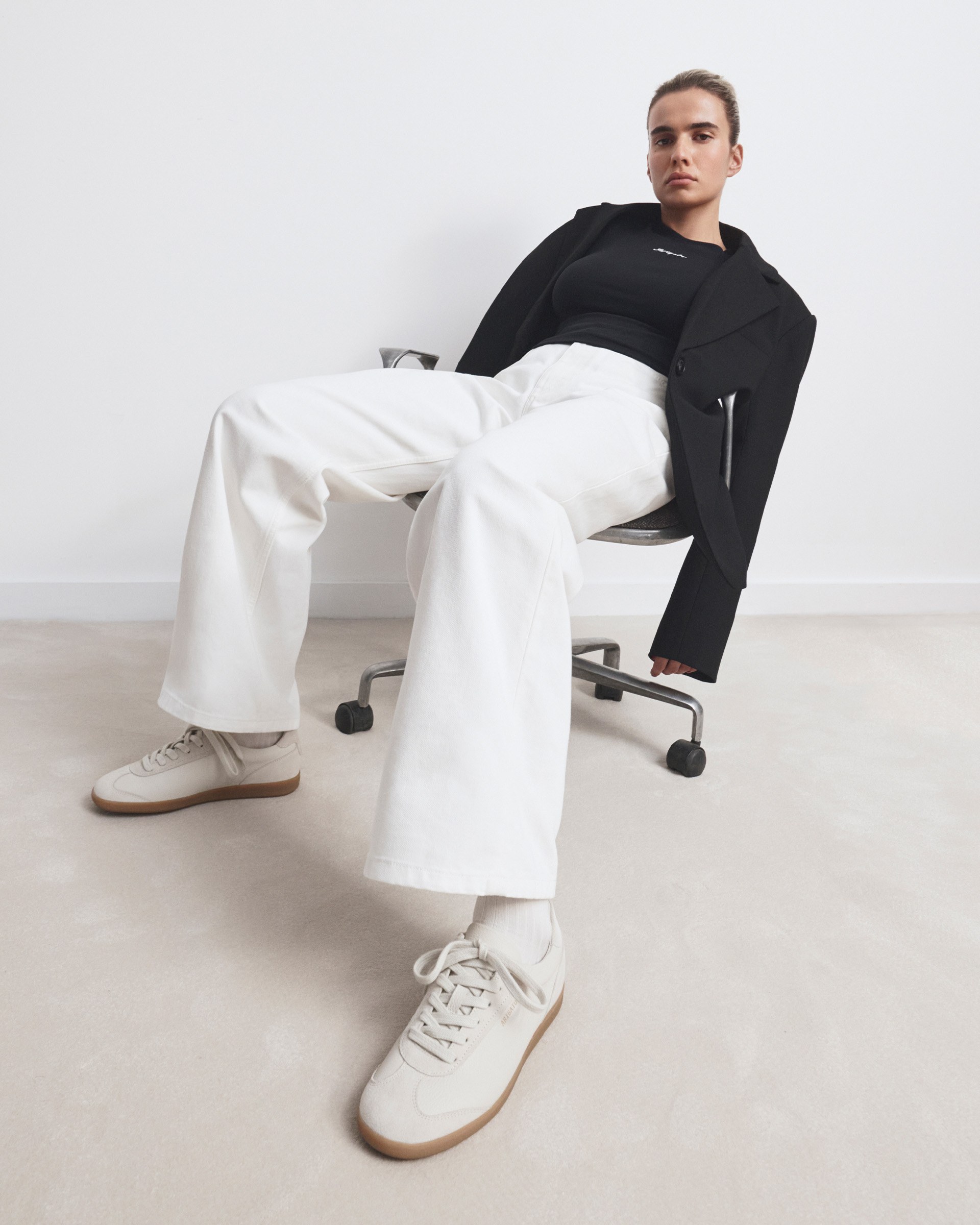 Person sitting on a chair, wearing a black top, black blazer, white trousers, and white sneakers, against a plain background.
