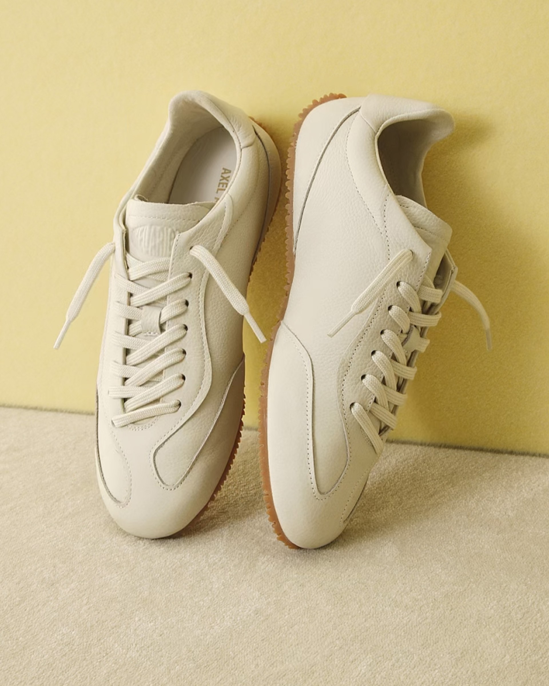 Pair of stylish, off-white leather sneakers with laces, positioned against a light yellow background and soft beige surface.