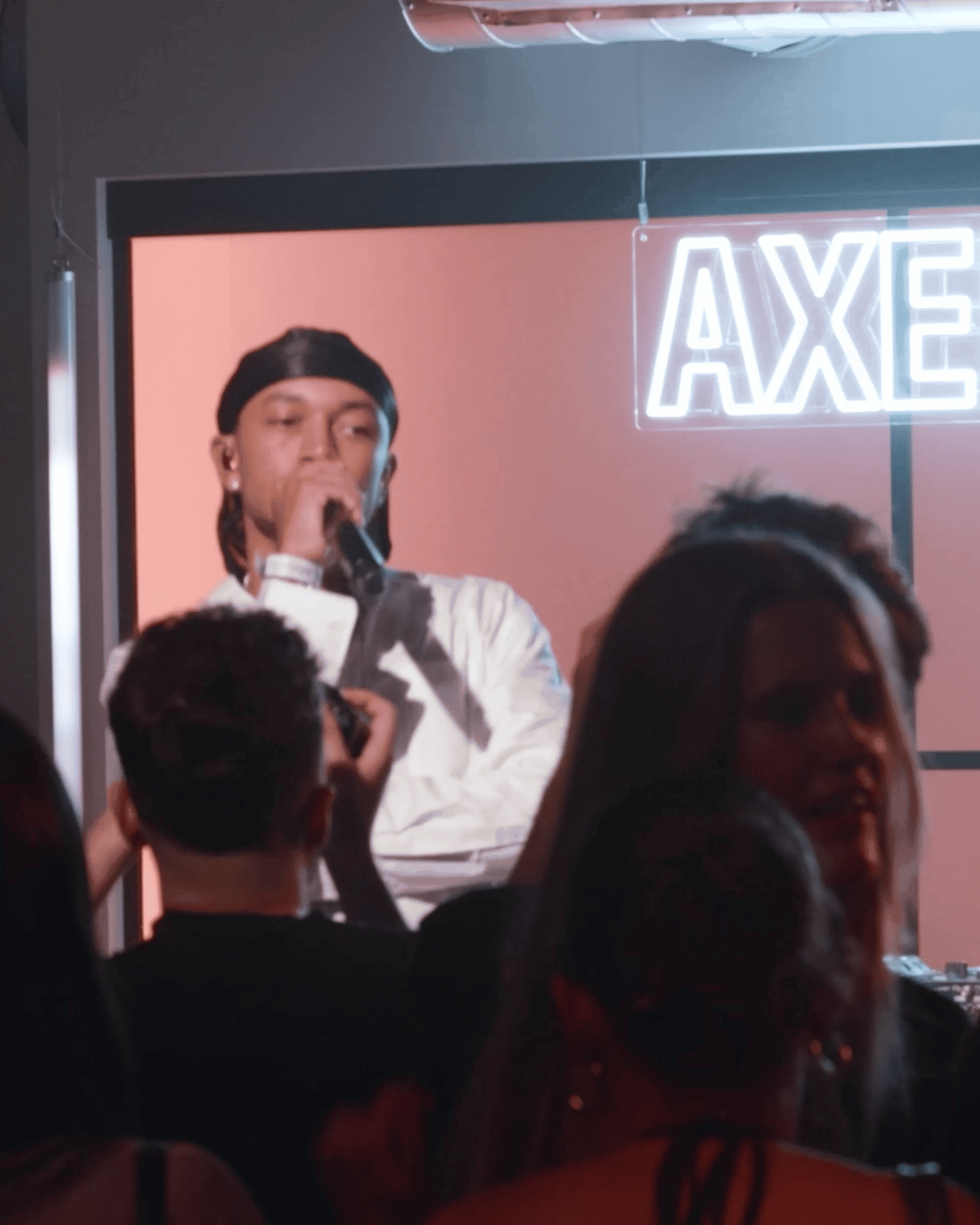 Performer on stage with microphone, wearing a durag, in front of an audience under a neon "AXE" sign.