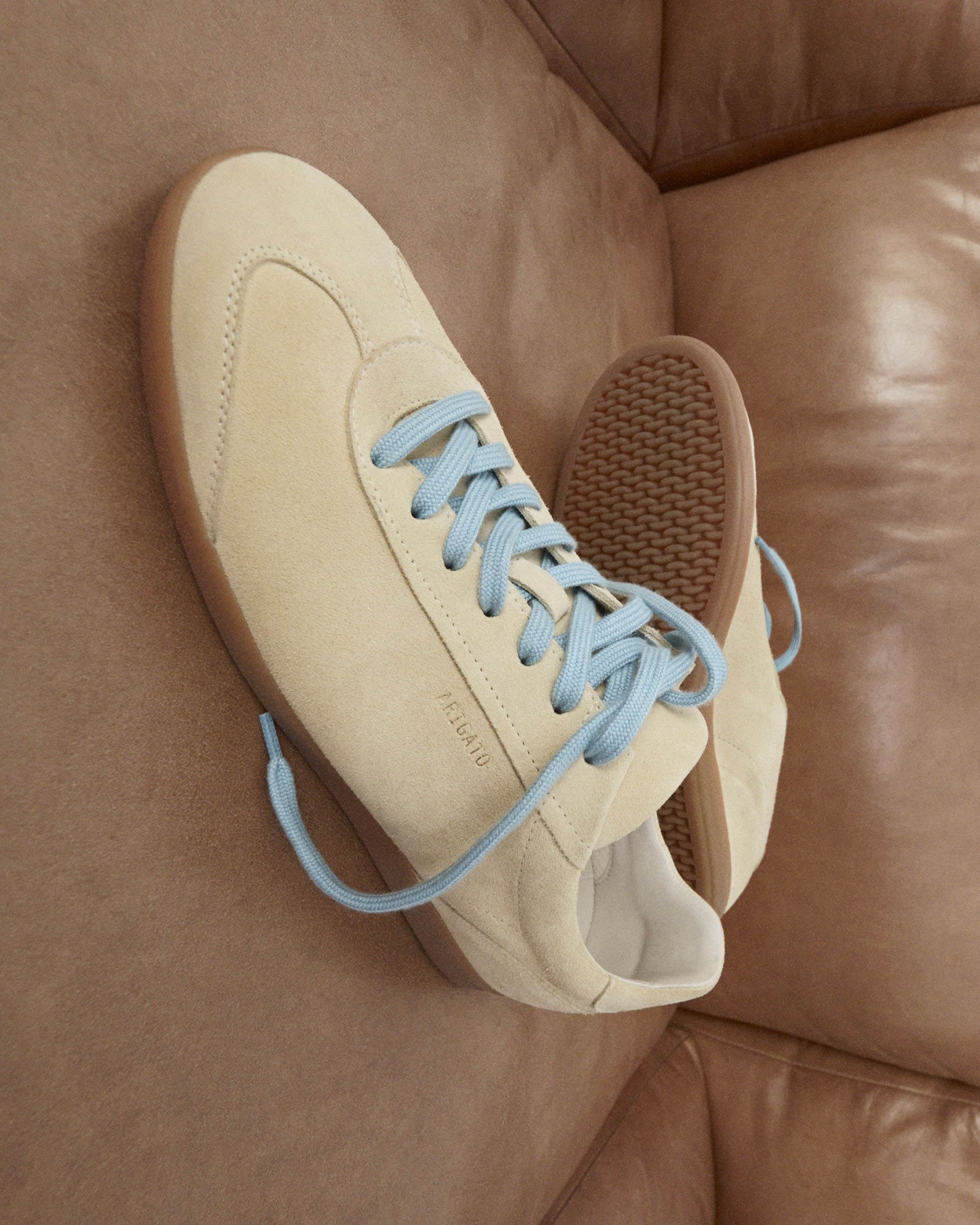 Beige suede sneakers with blue laces on a brown leather couch, showing side and top views.