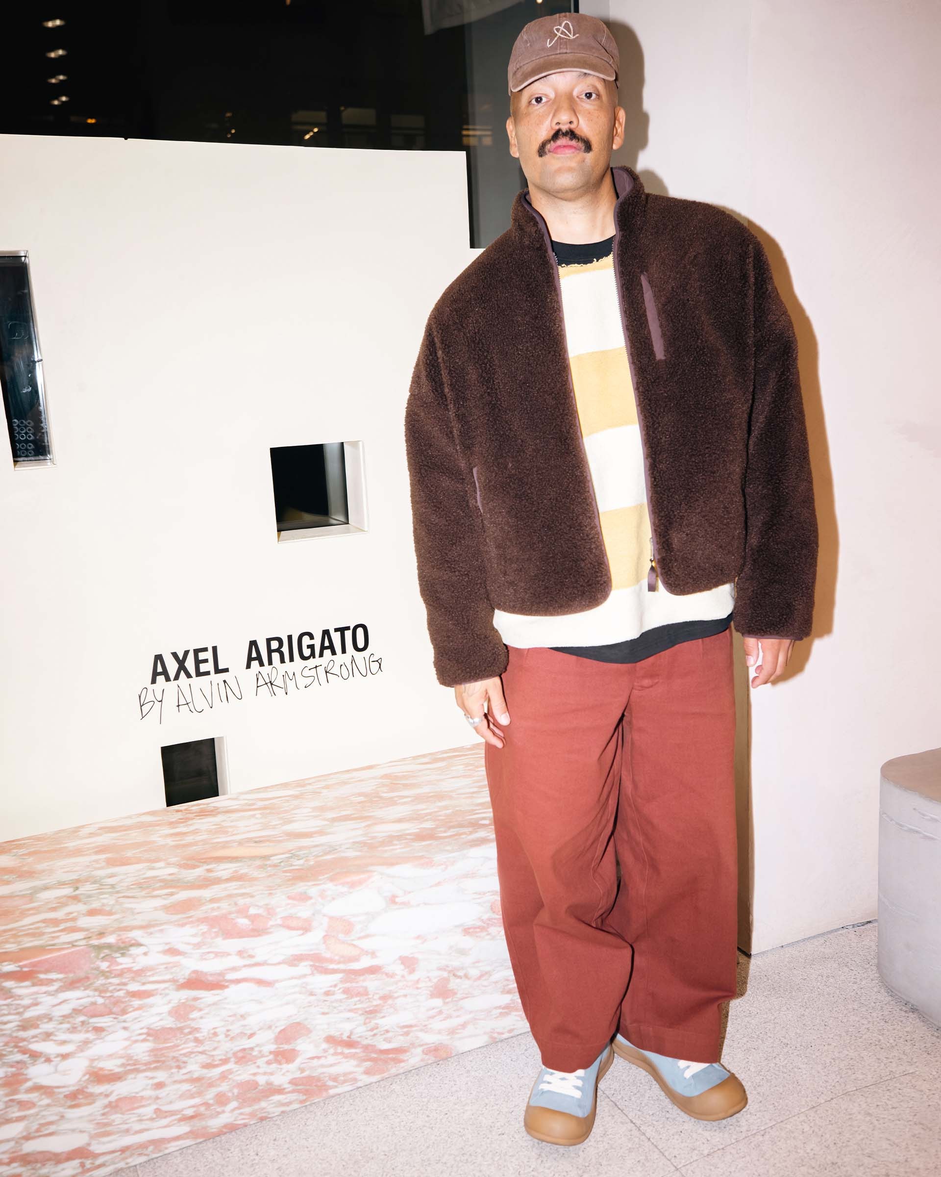 Person in a brown jacket and red pants stands indoors against a white wall with "Axel Arigato" text.
