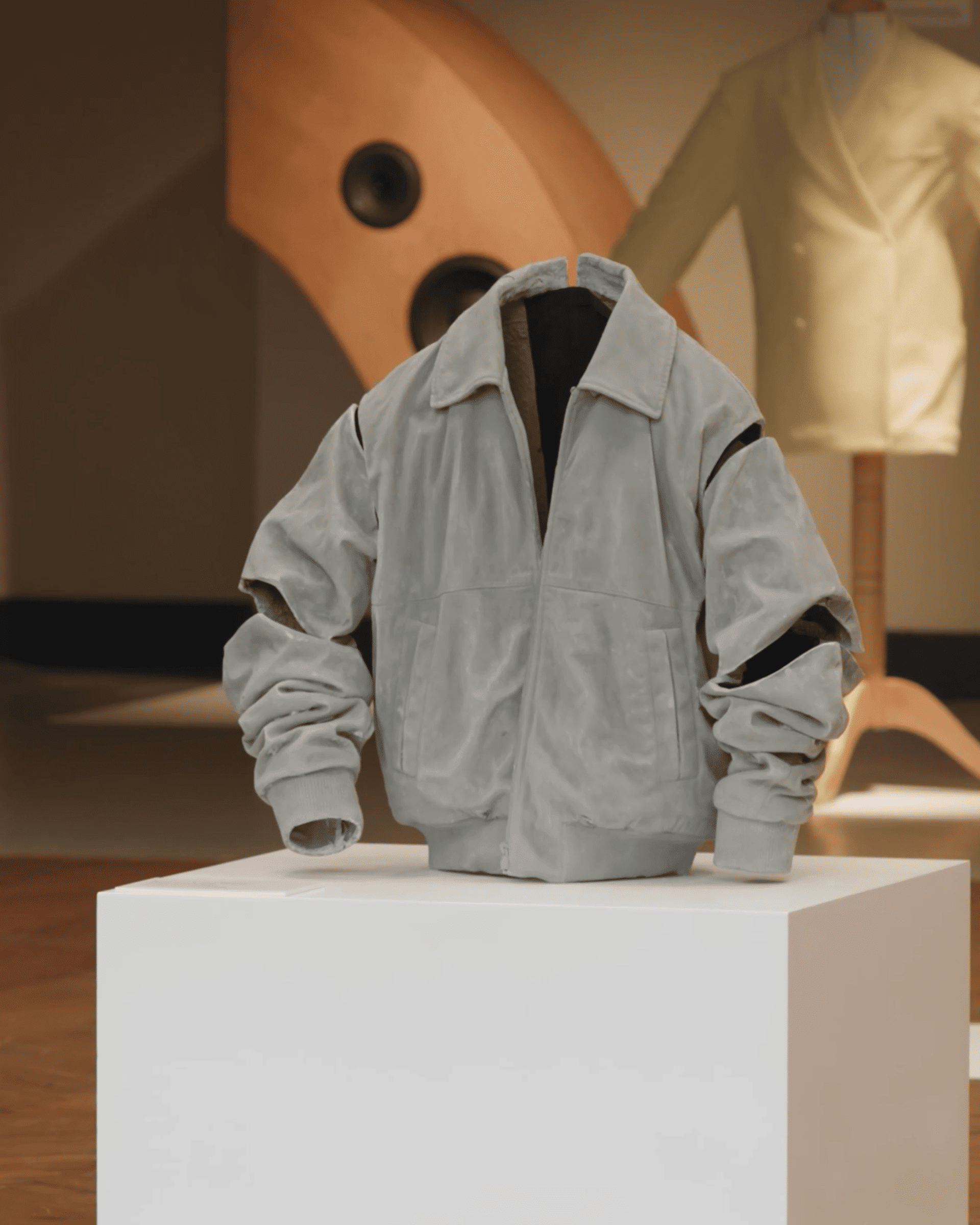 A light gray bomber jacket displayed on a white pedestal in a gallery setting, with a blurred background featuring abstract shapes and a mannequin.