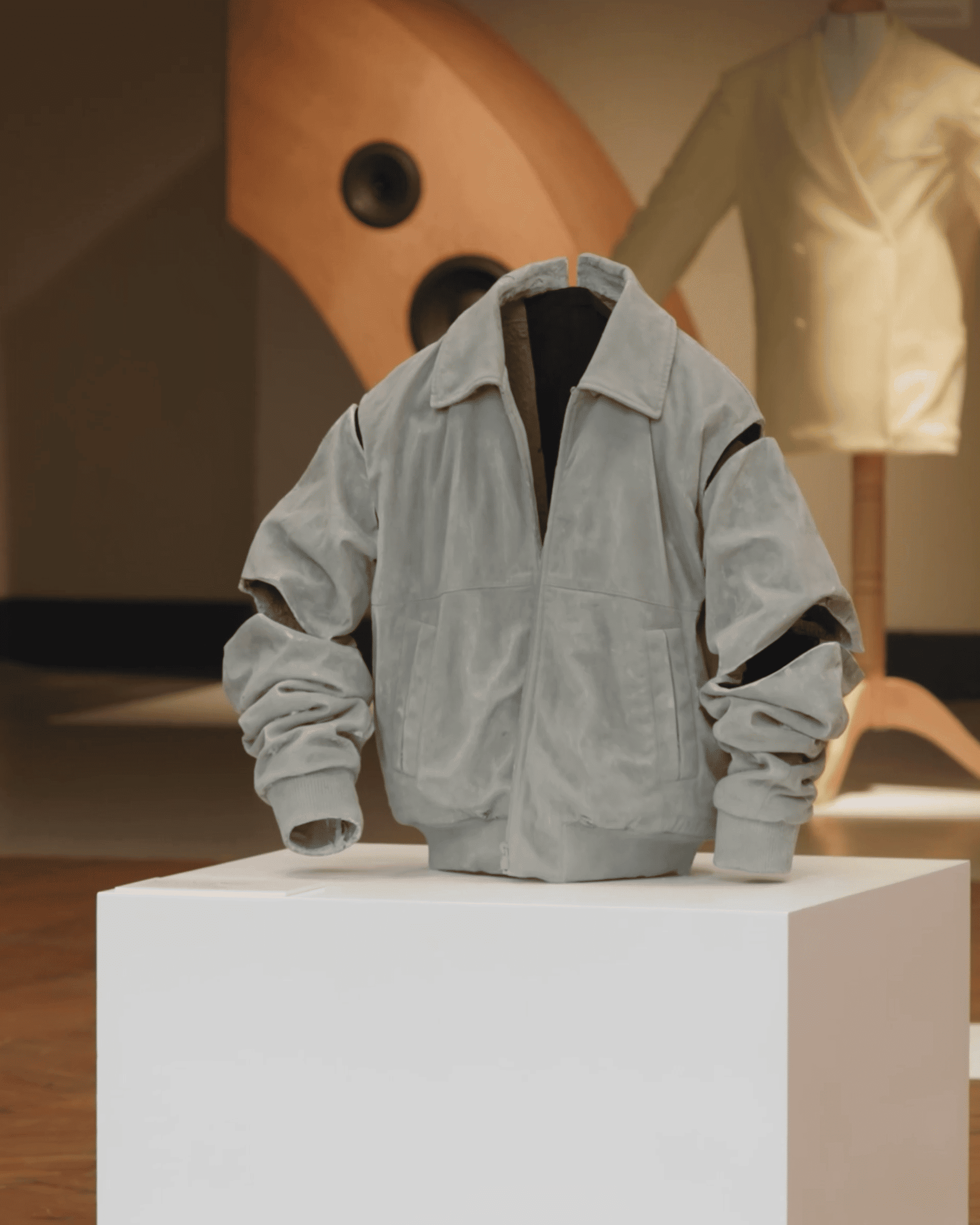 A light gray bomber jacket displayed on a white pedestal in a gallery setting, with a blurred background featuring abstract shapes and a mannequin.