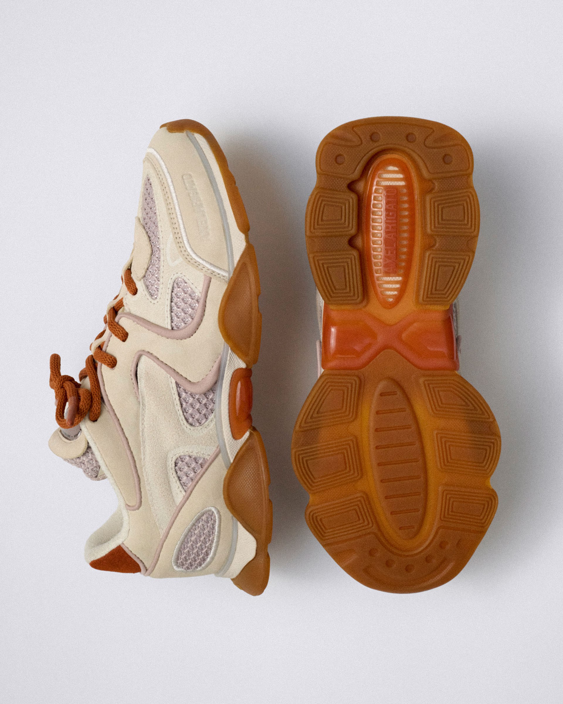 A pair of beige and orange sneakers, one showing the side with intricate mesh designs, the other displaying the textured rubber sole.