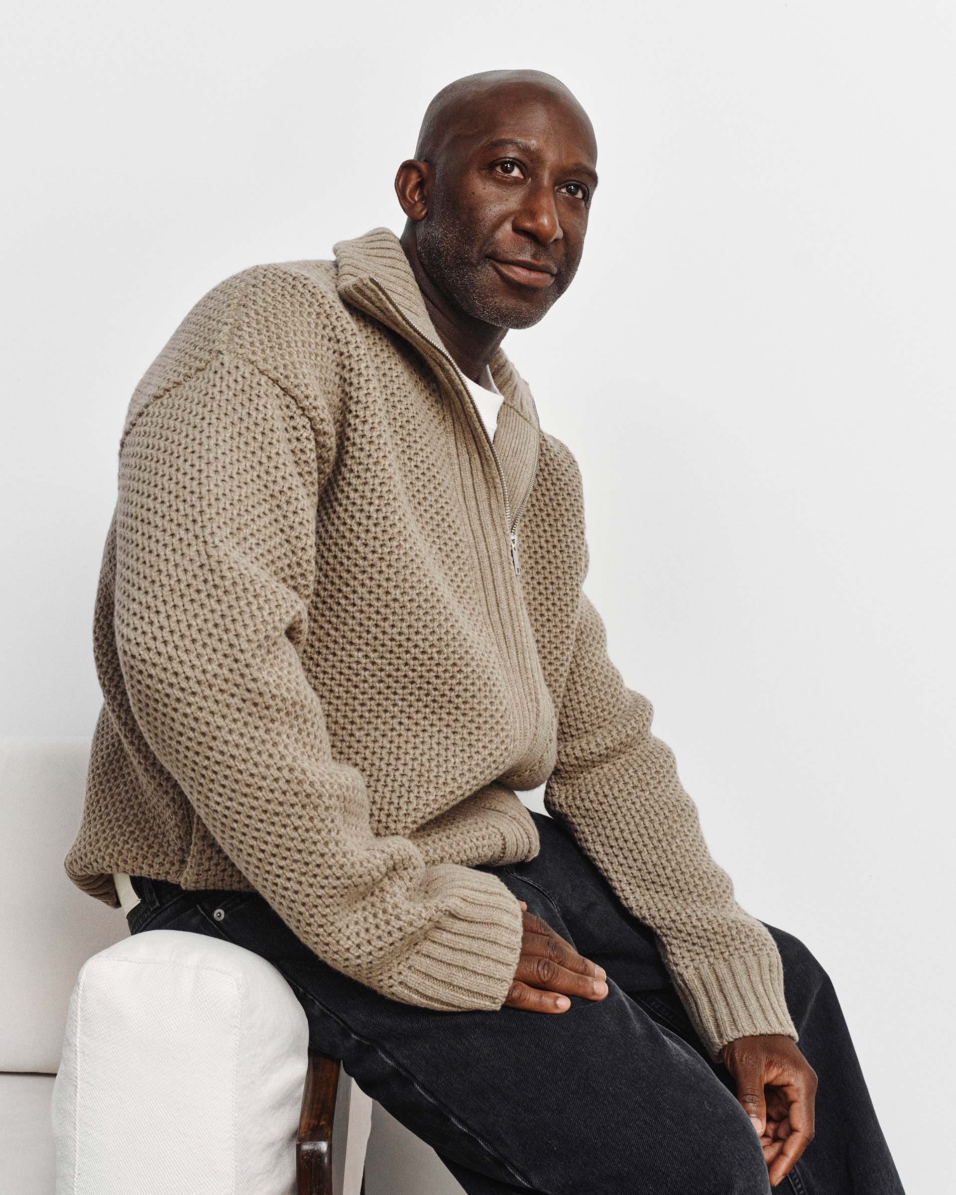 Man sitting on a white chair, wearing a beige knit sweater and dark pants, looking thoughtfully to the side against a plain background.