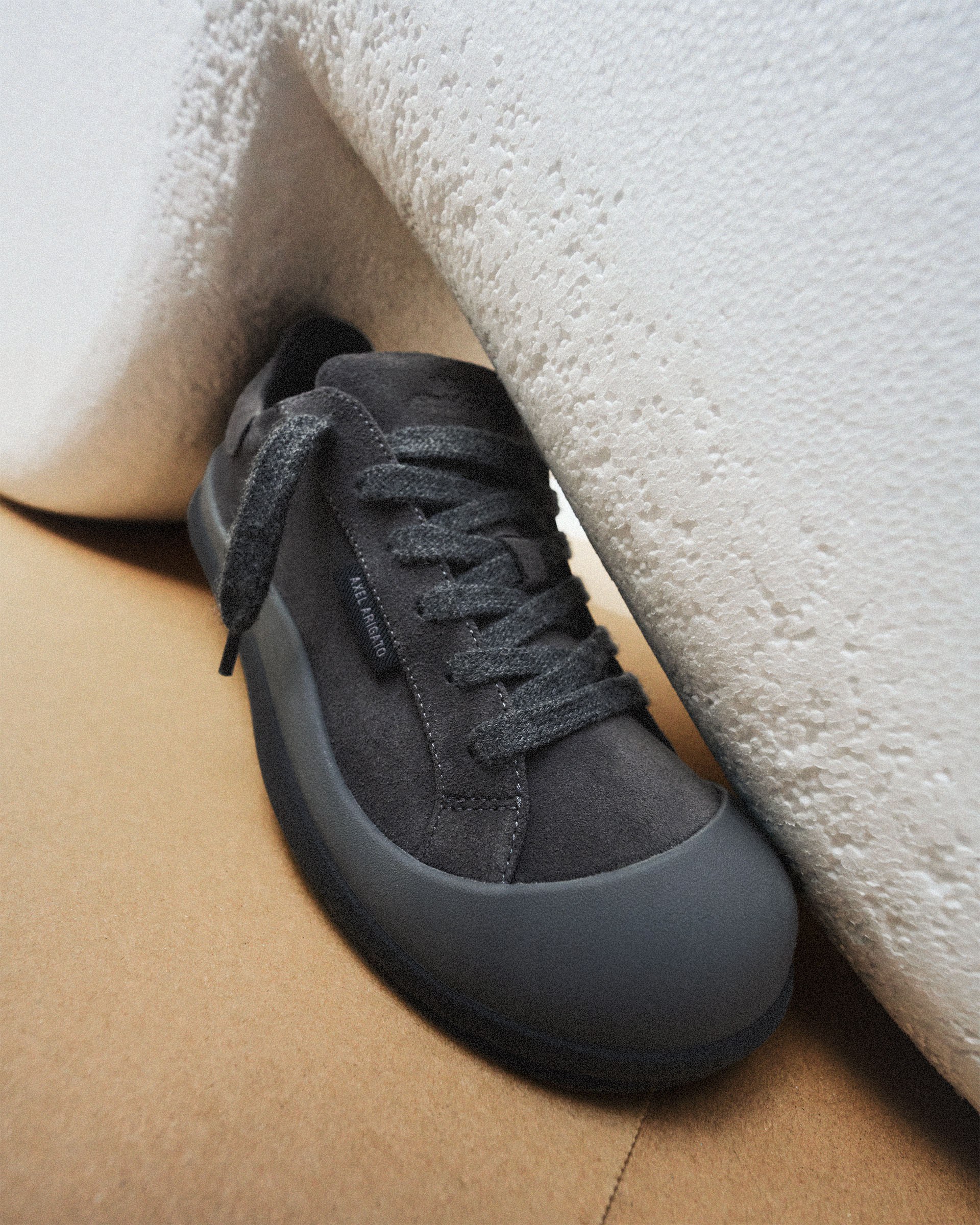 Dark gray sneaker with black laces resting against a textured white surface on a light brown background.
