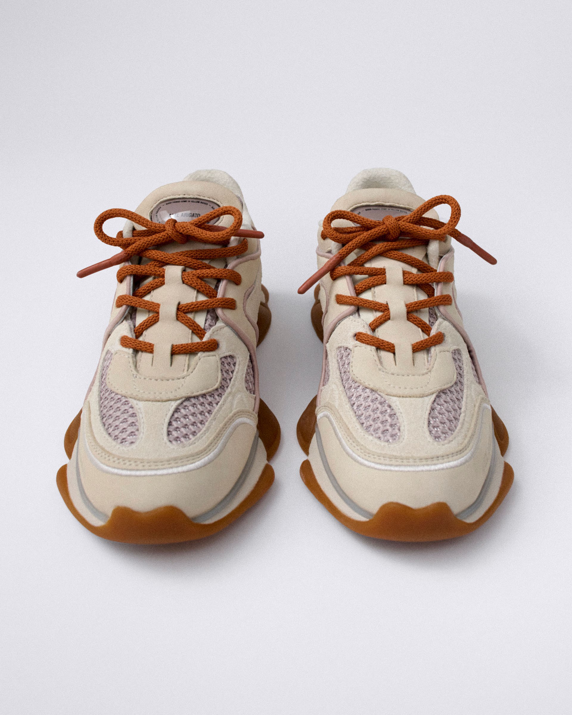 A pair of cream and tan sneakers with orange laces, featuring a wavy sole and mesh detailing on the upper, displayed on a neutral background.