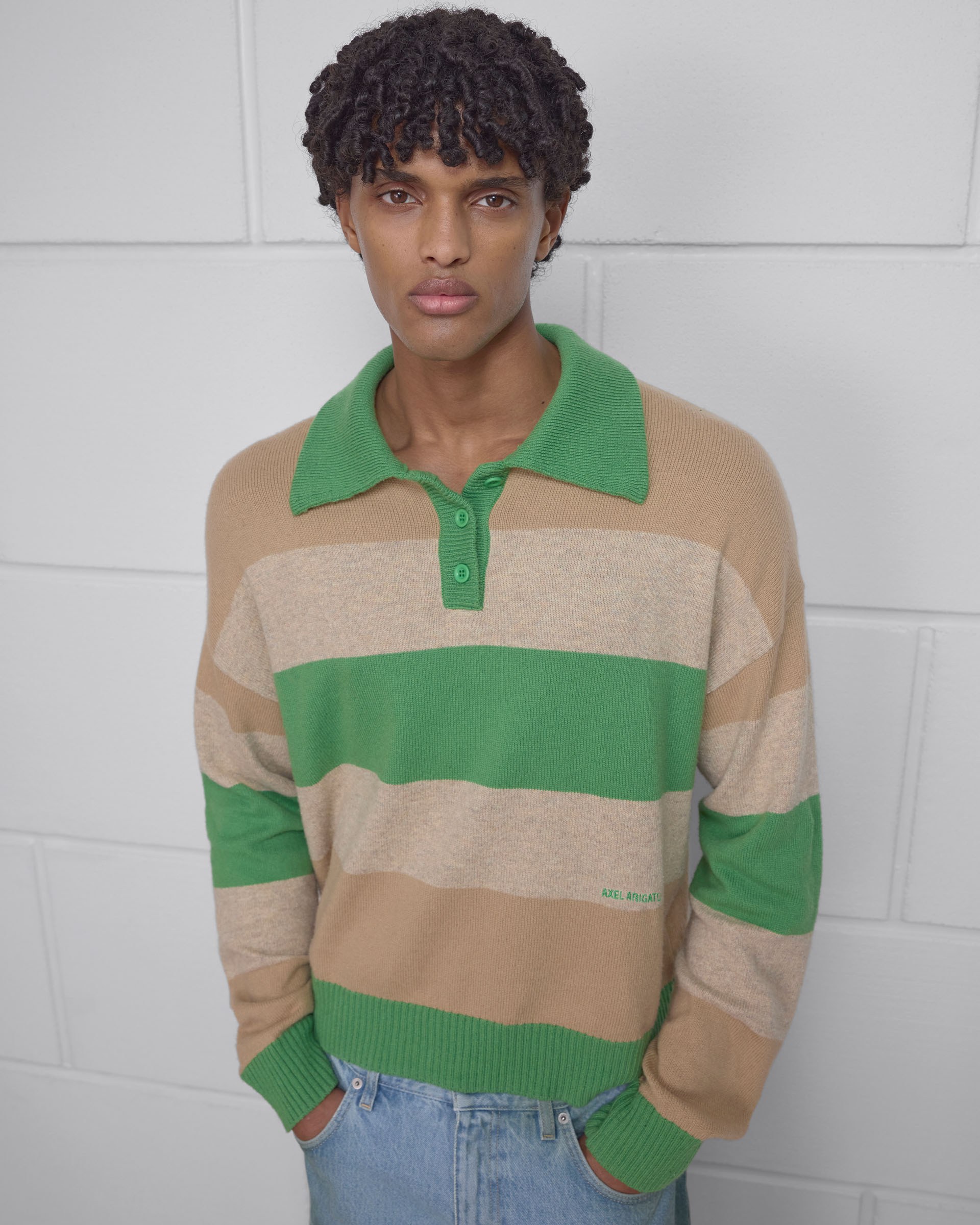 Person wearing a green and beige striped sweater with a green collar, standing against a white brick wall.