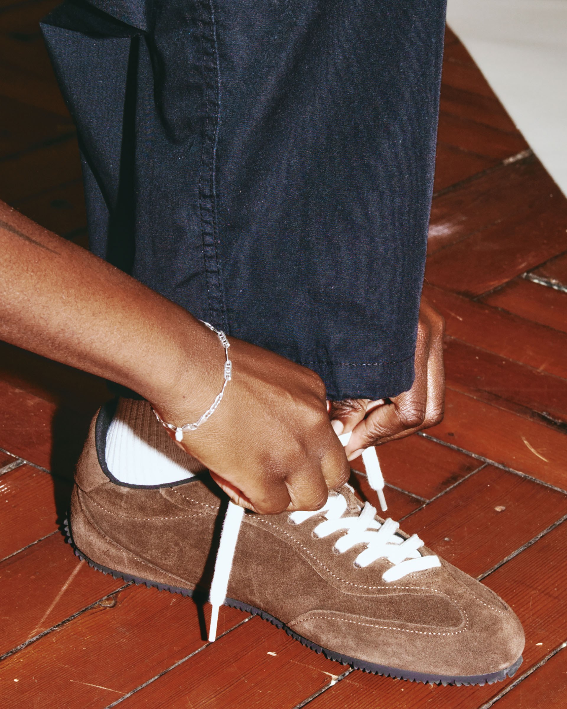 Person tying the laces of brown suede sneakers, wearing black pants and a silver bracelet, on a wooden floor.