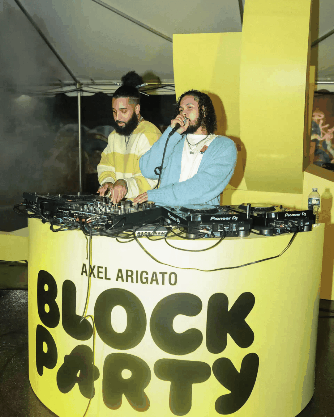 Two DJs perform at the Axel Arigato Block Party, with one speaking into a microphone behind a yellow DJ booth setup.