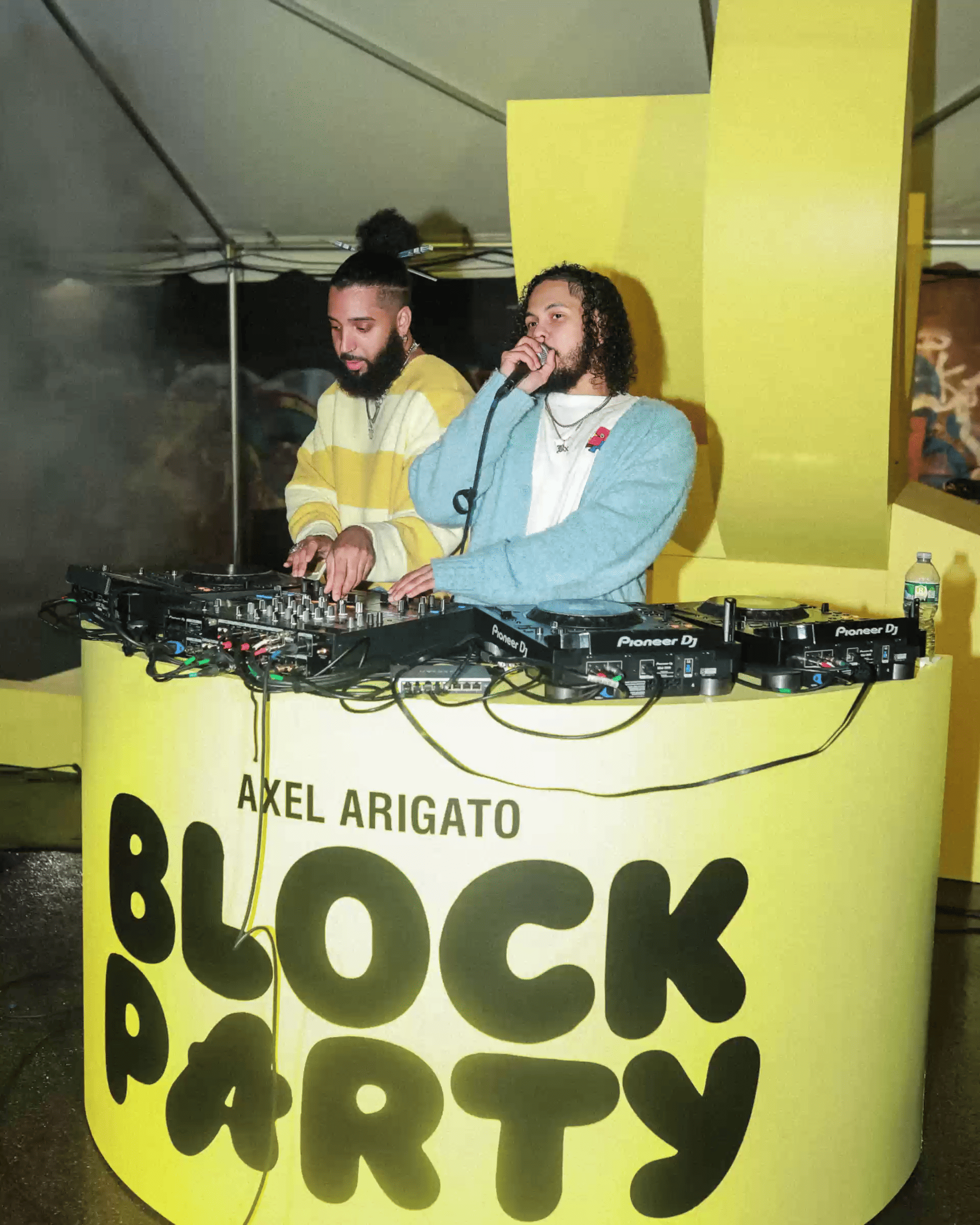 Two DJs perform at the Axel Arigato Block Party, with one speaking into a microphone behind a yellow DJ booth setup.