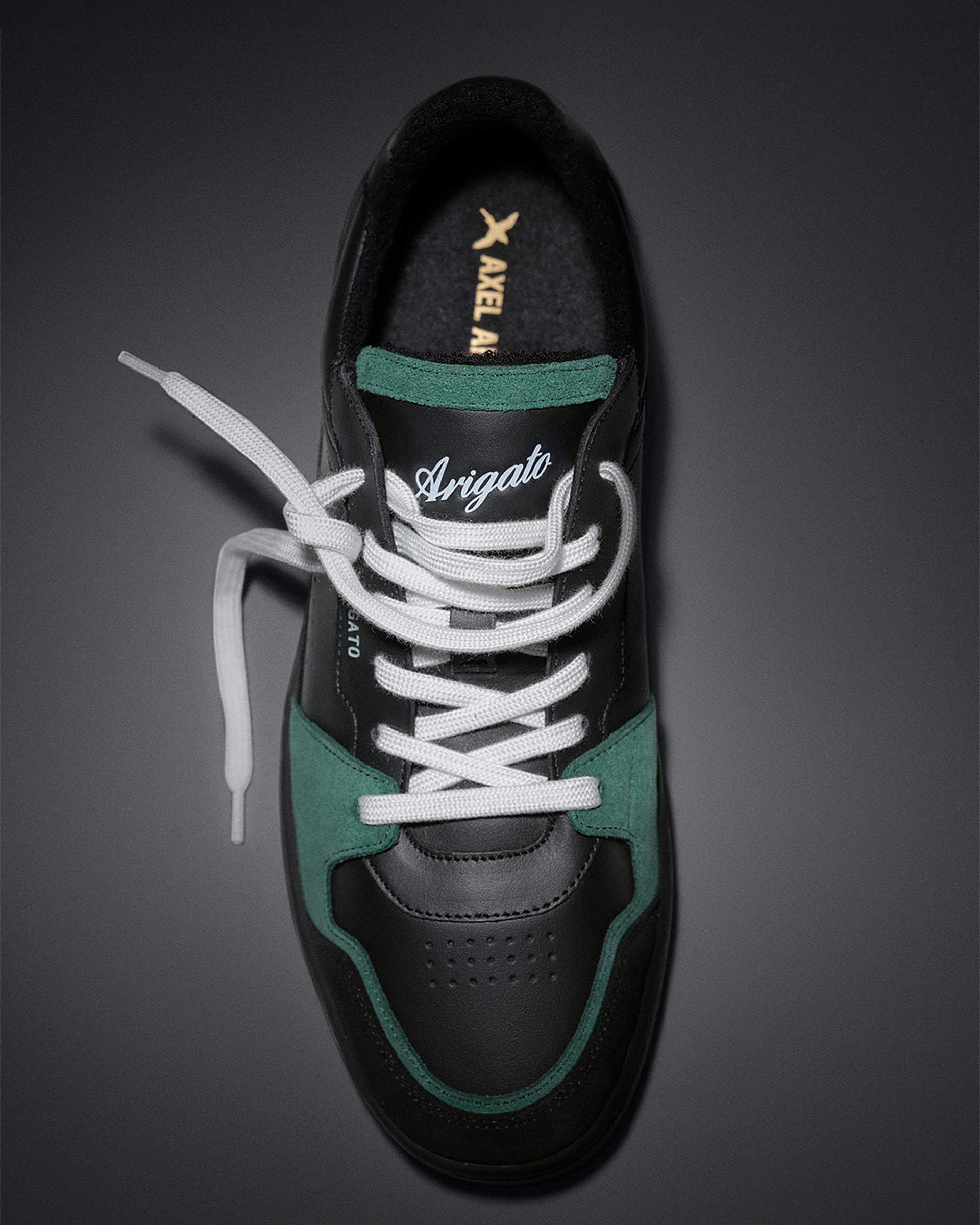 Black and green sneaker with white laces and "Arigato" written on the tongue, set against a dark background.