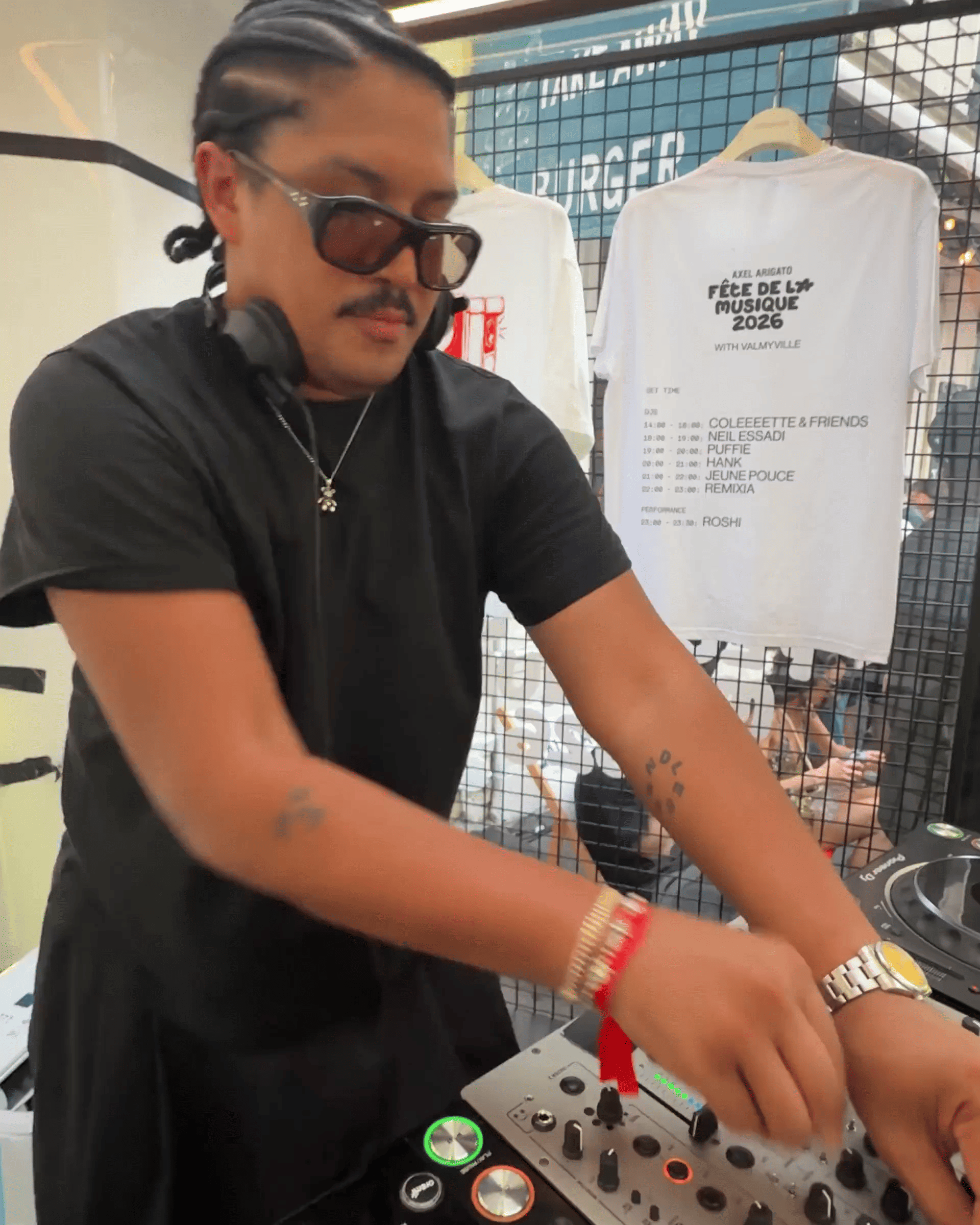 DJ with braided hair and sunglasses mixing music on a set of turntables, a T-shirt hanging in the background.