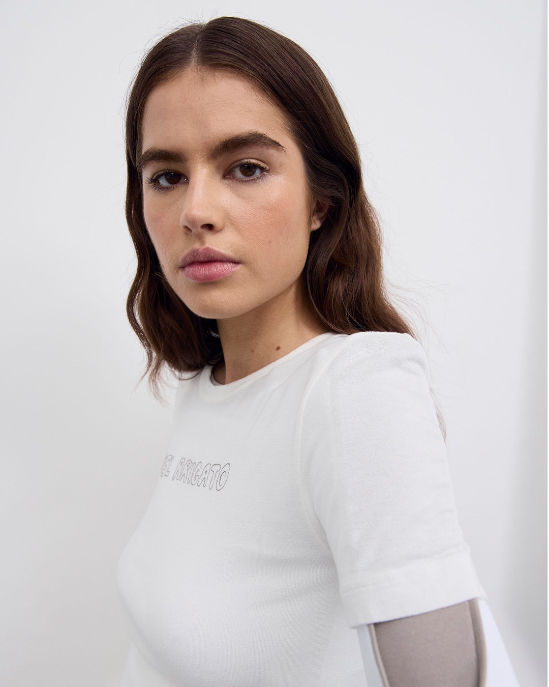 A woman with long brown hair wearing a white shirt looks directly at the camera against a plain background.