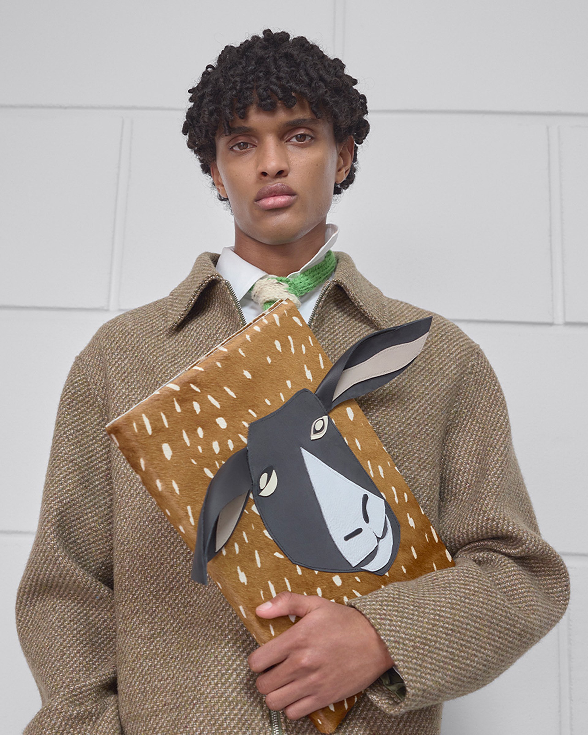 A person in a brown coat holds a clutch bag with a stylized sheep face design, against a white tiled wall.