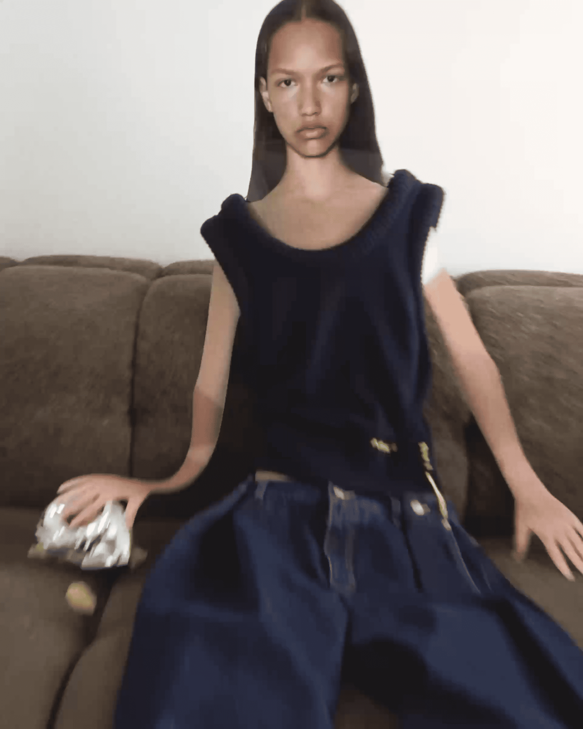 Person sitting on a couch wearing a dark sleeveless top and loose pants, with a blurred effect and a snack bag beside them.