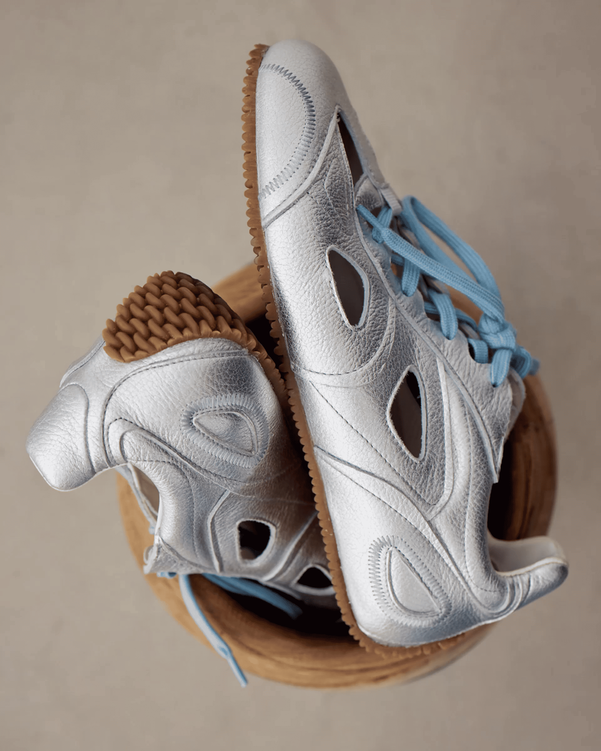 Silver athletic shoes with light blue laces and textured soles, creatively positioned in a wooden bowl on a beige background.