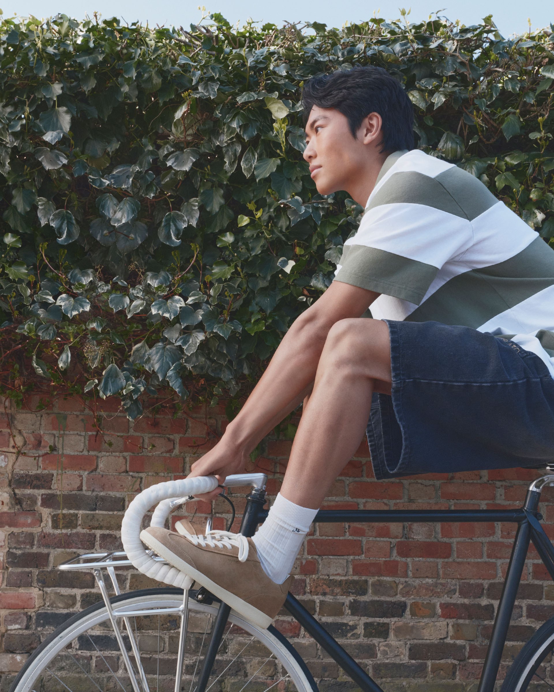 Man in striped shirt and denim shorts sits casually on a stationary bike. Brick wall and ivy background.