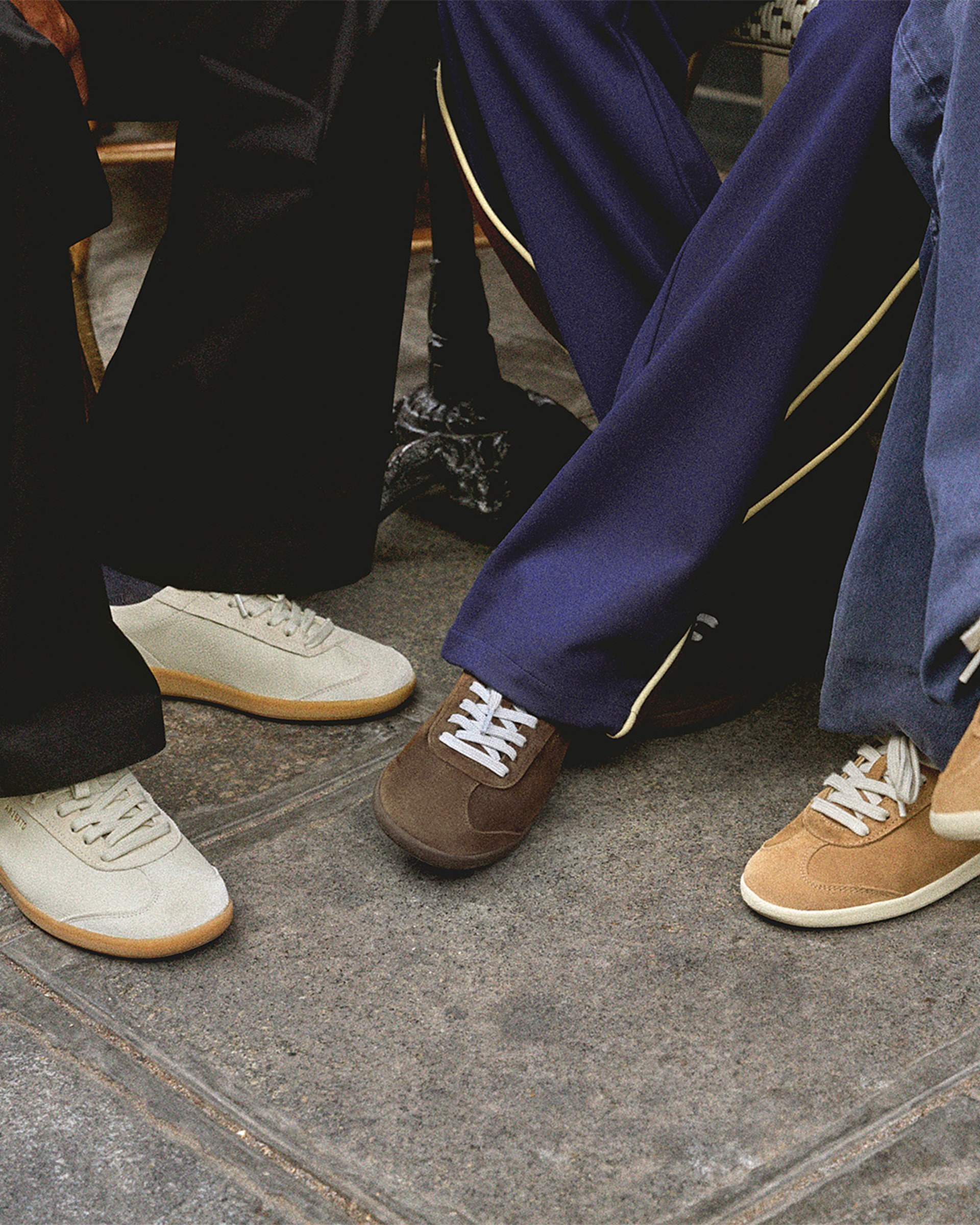 Three people wearing different colored sneakers and flared pants stand on a stone pavement, showing off their footwear styles.