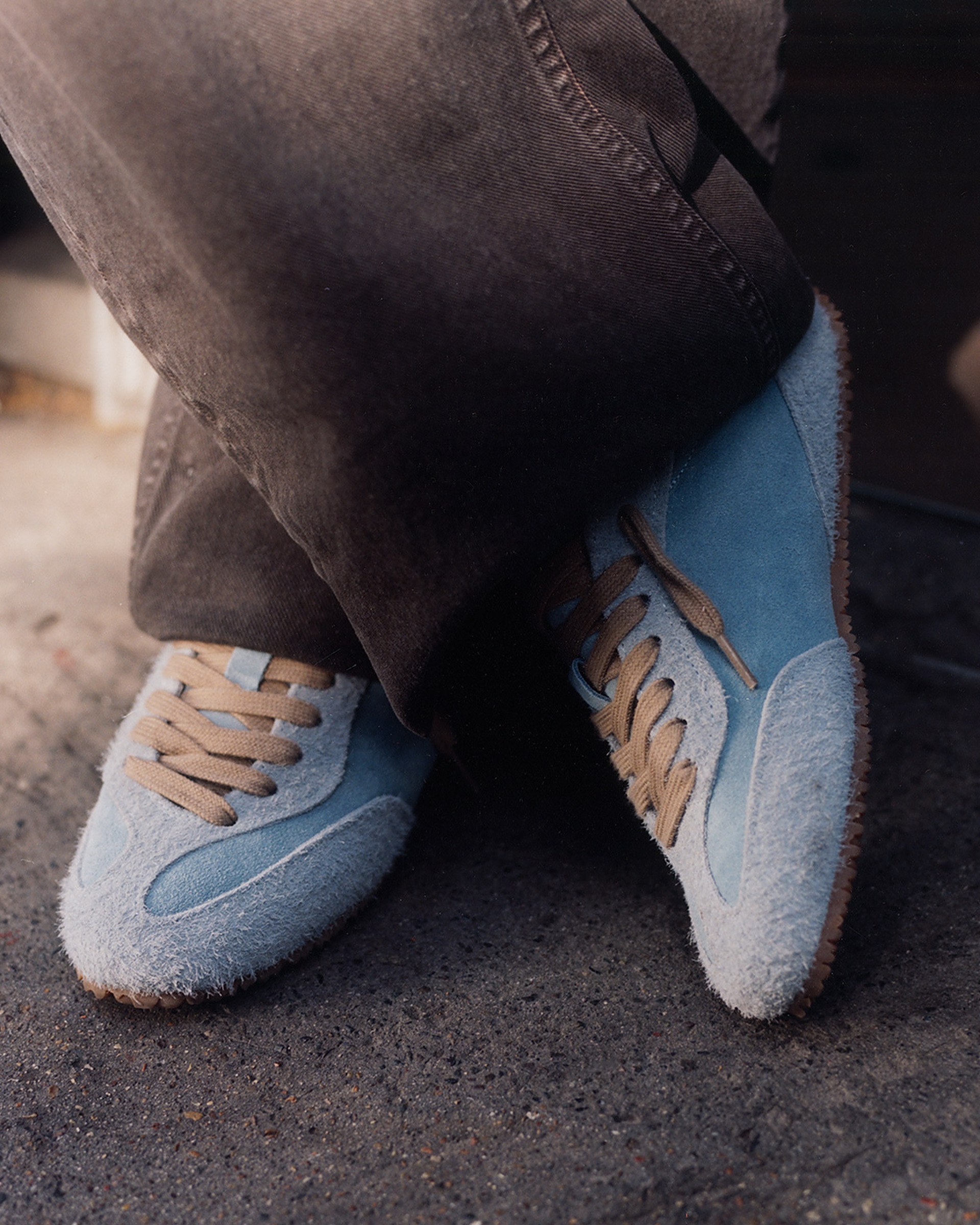 Person wearing light blue suede sneakers with tan laces and gum soles, paired with dark brown pants, standing on a concrete surface.
