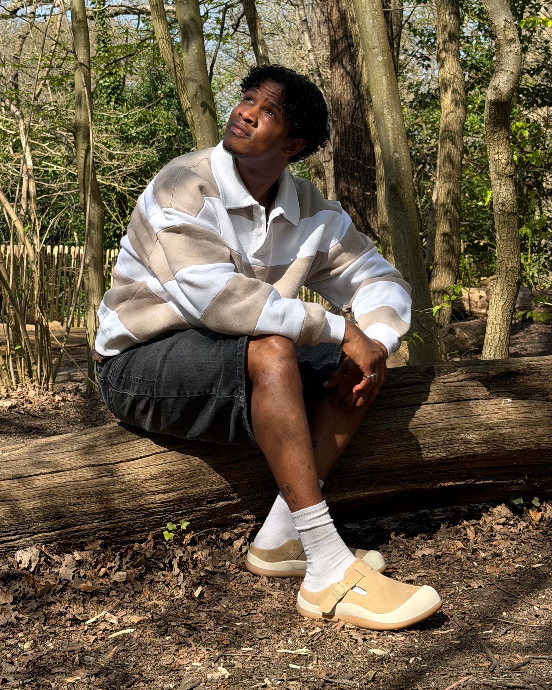 Person sitting on a log in a forest, wearing a striped shirt, black shorts, socks, and beige shoes, looking up thoughtfully.
