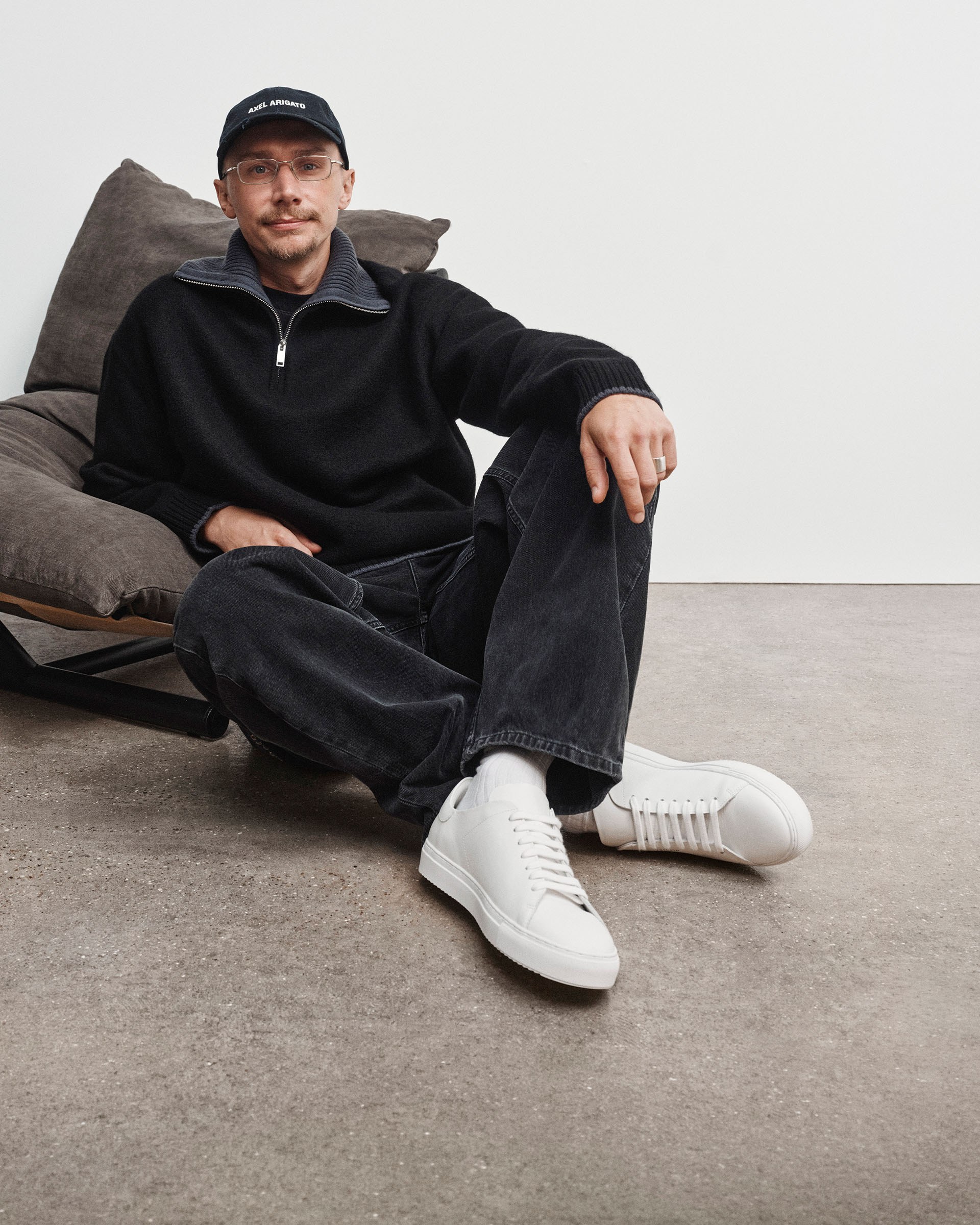A man in a black cap and sweater sits on the floor against a chair, wearing dark jeans and white sneakers, with a relaxed expression.