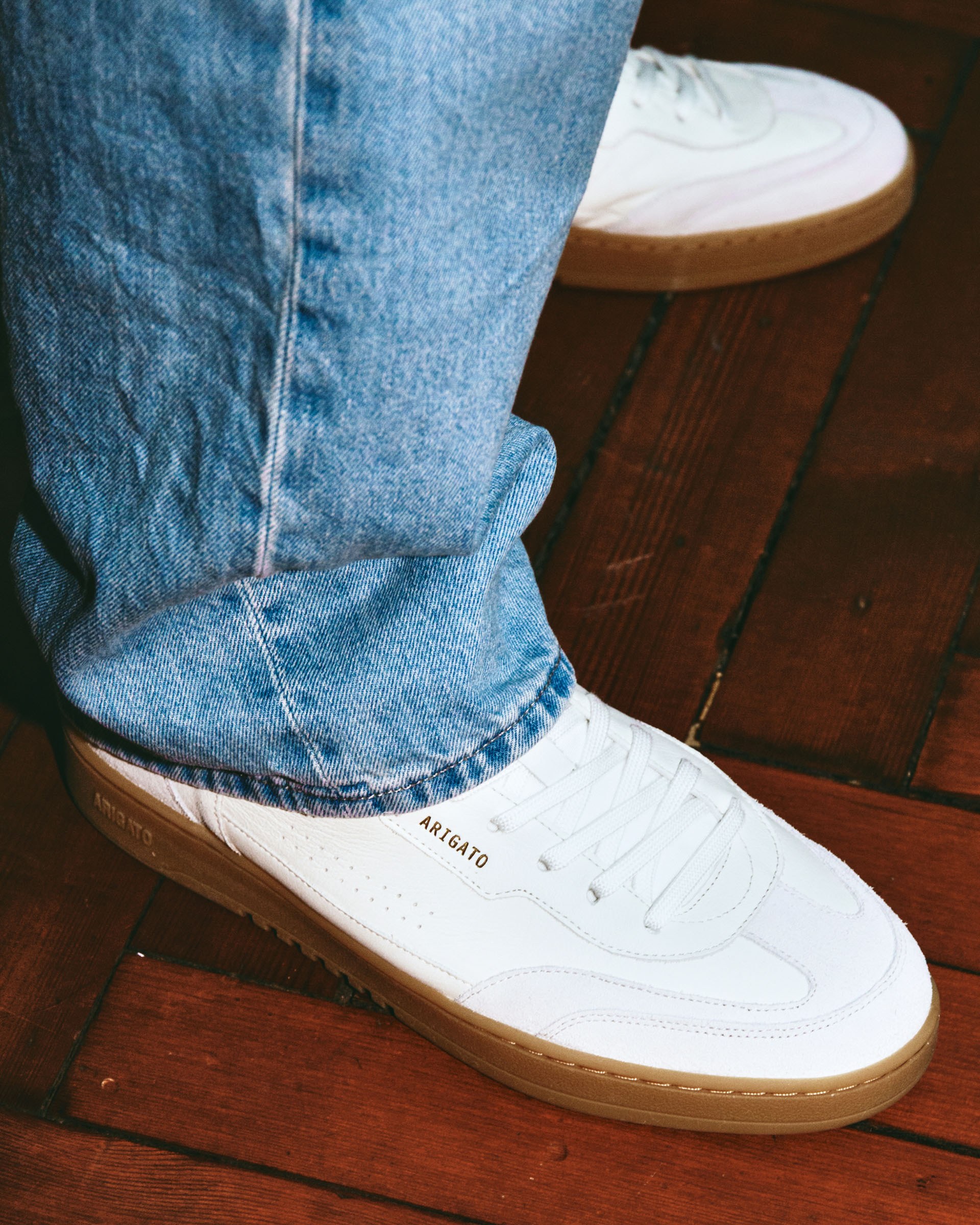 White sneakers with gum soles and "Arigato" branding, worn with light blue jeans, on a wooden floor.