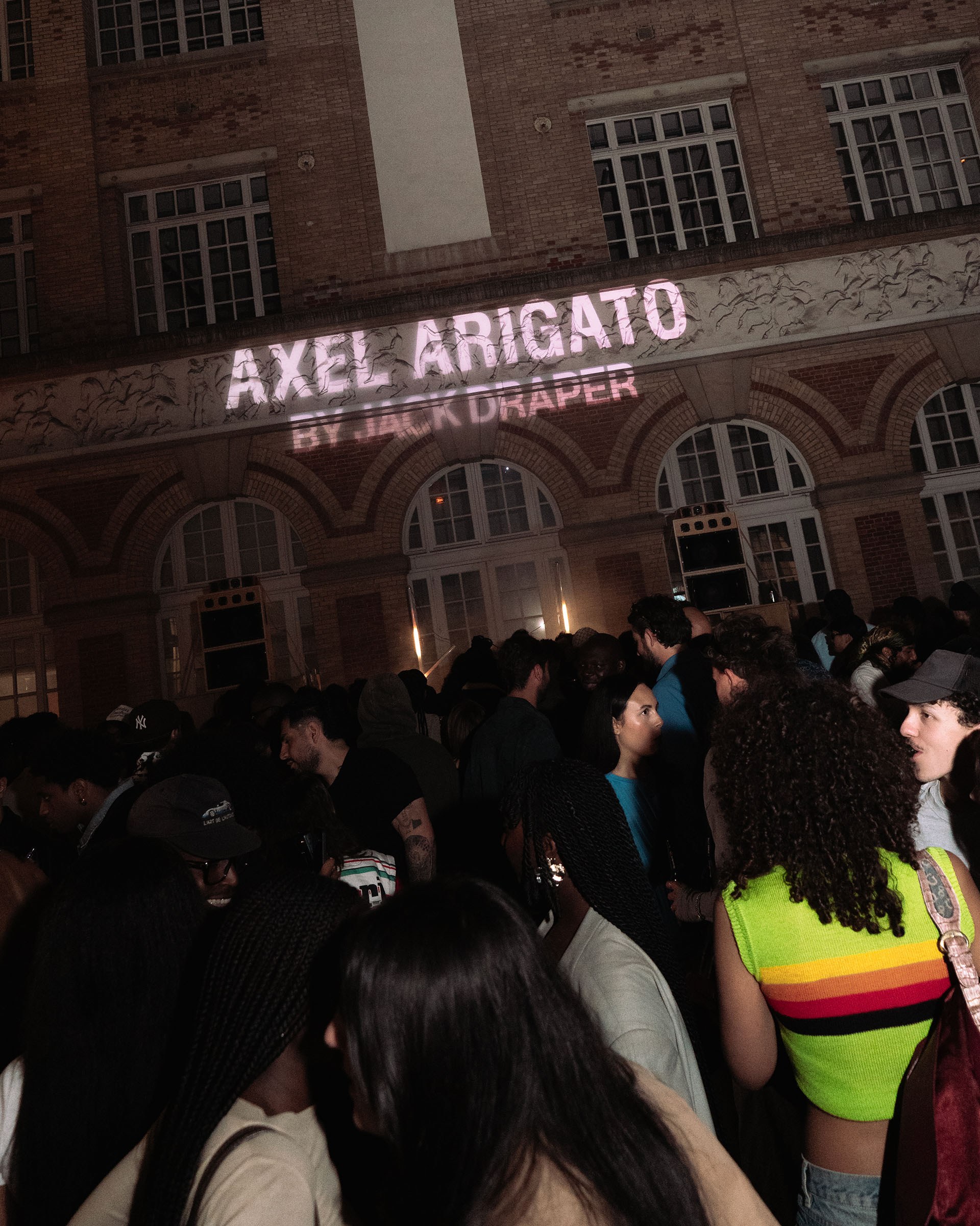 A lively crowd attends an evening event at a building with "Axel Arigato by Jack Draper" projected on the façade.
