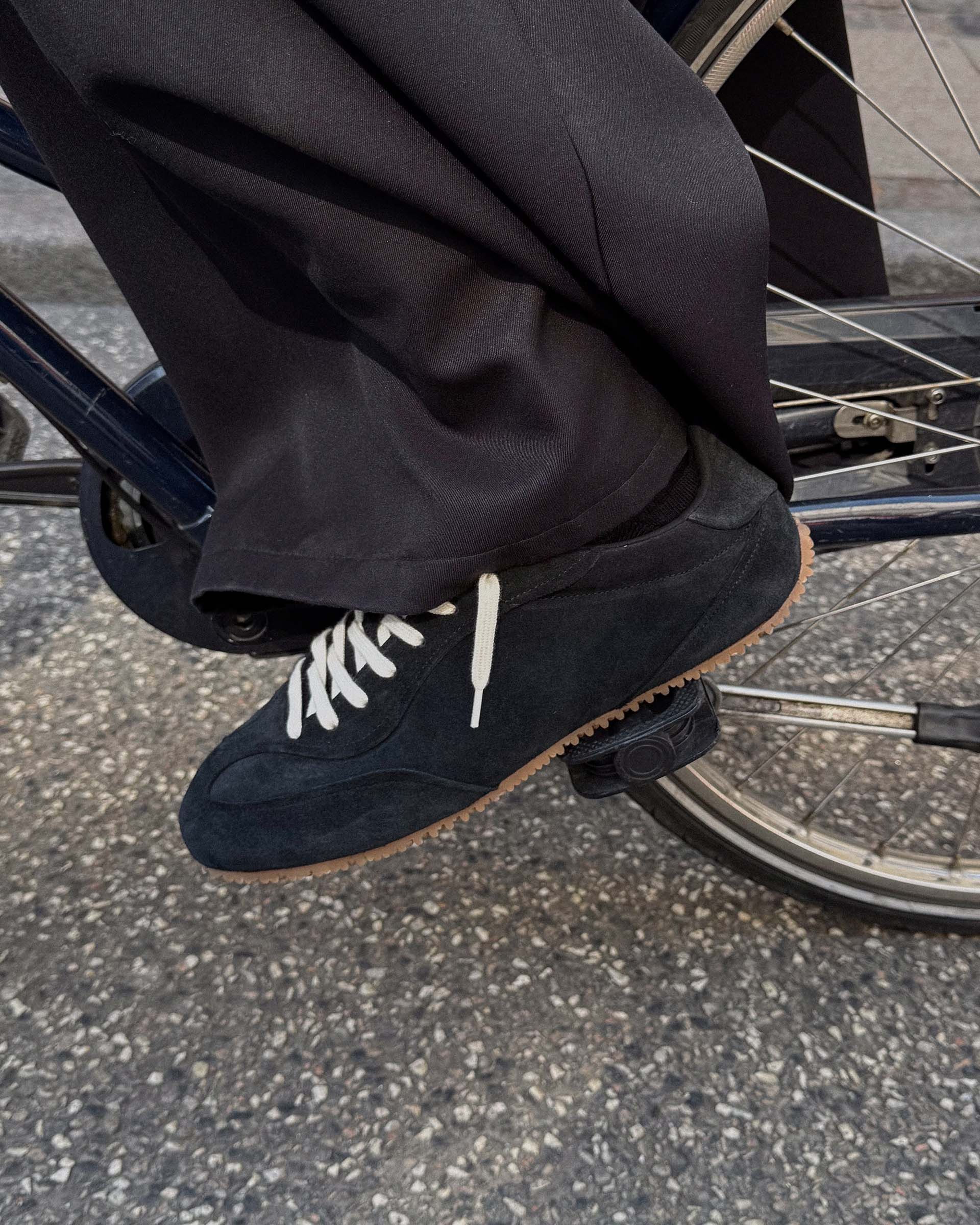 Person wearing black pants and dark sneakers with white laces, riding a bicycle on a paved road.