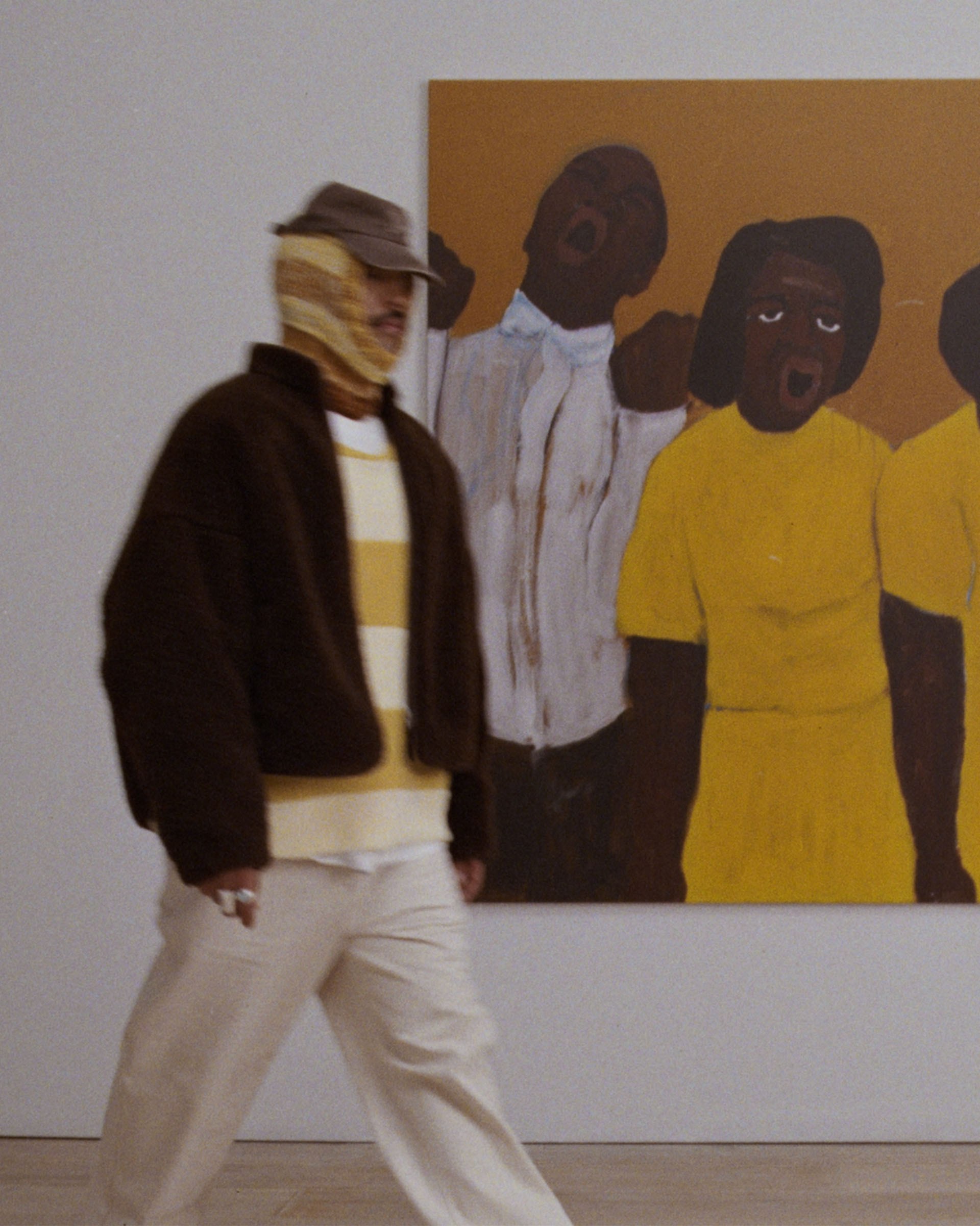 Person in layered clothing walks past a painting of people in yellow and white outfits on a gallery wall.
