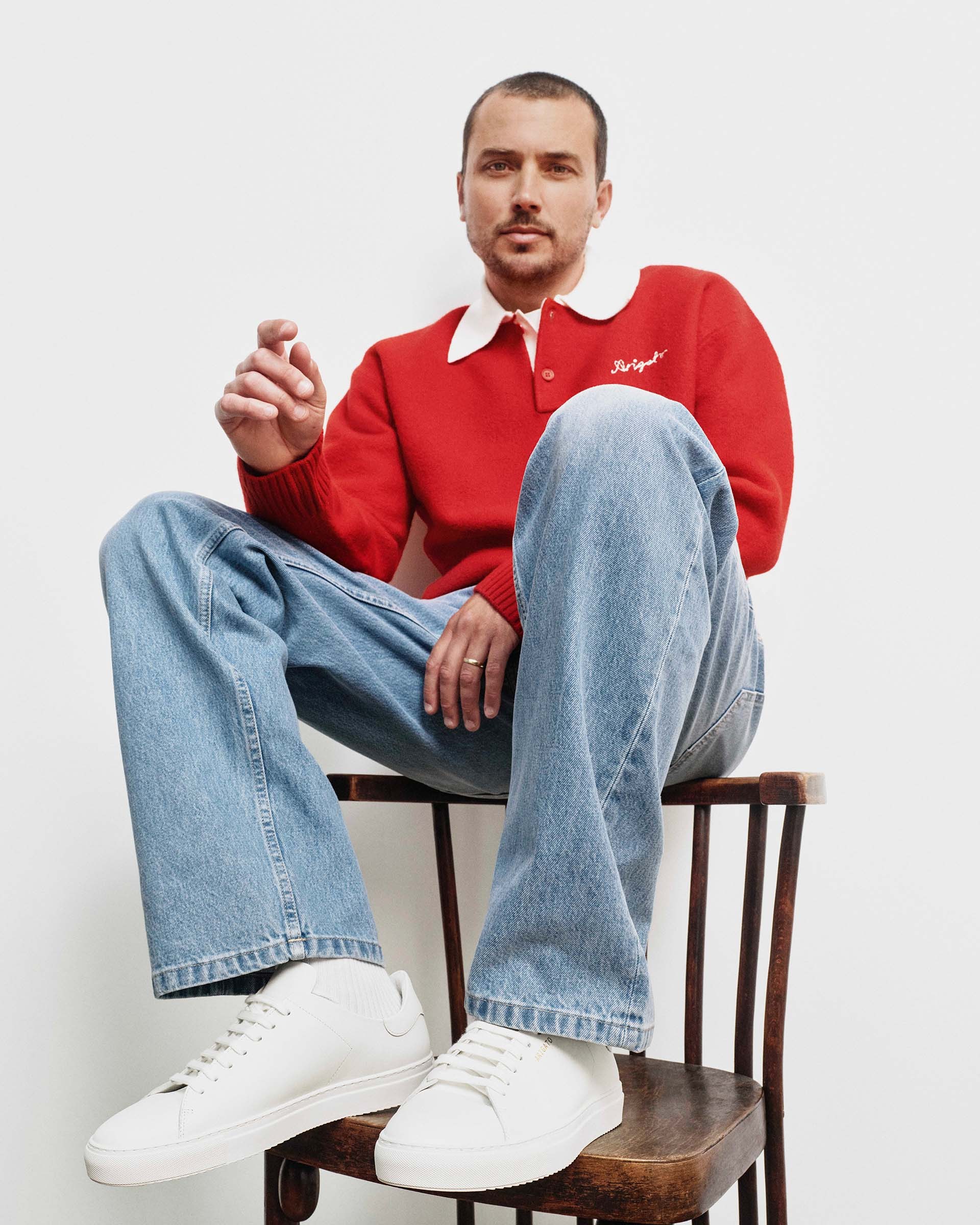 A man sits casually on a chair, wearing a red collared sweater, light blue jeans, and white sneakers, against a plain white background.