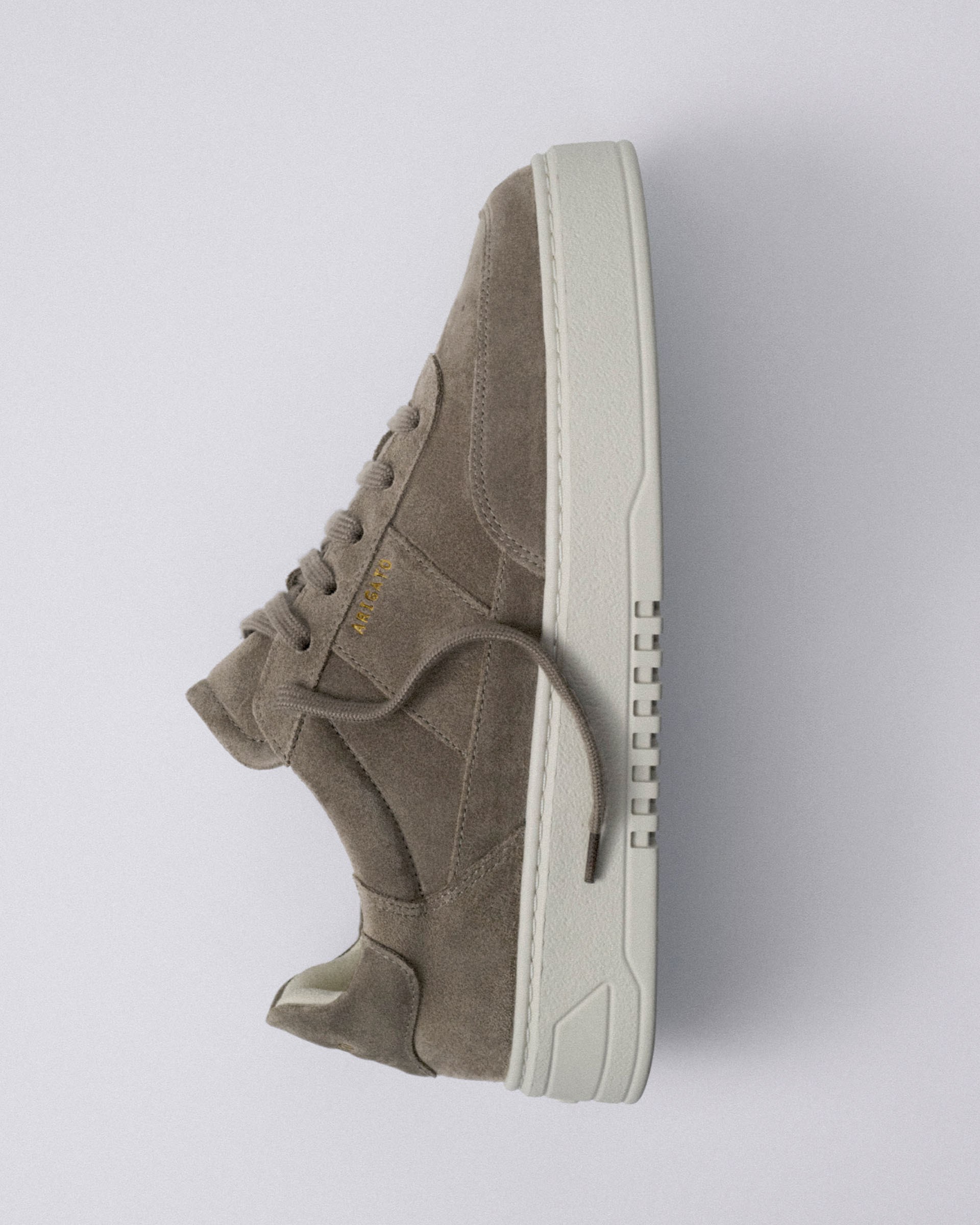 Brown suede sneaker with a white sole, side view. The shoe features a minimalist design with laces and subtle stitching details.