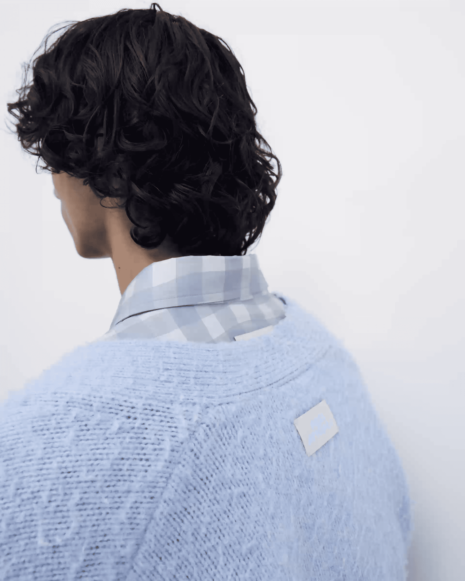 Person with curly hair wearing a plaid shirt under a soft, light blue sweater, facing away against a plain background.