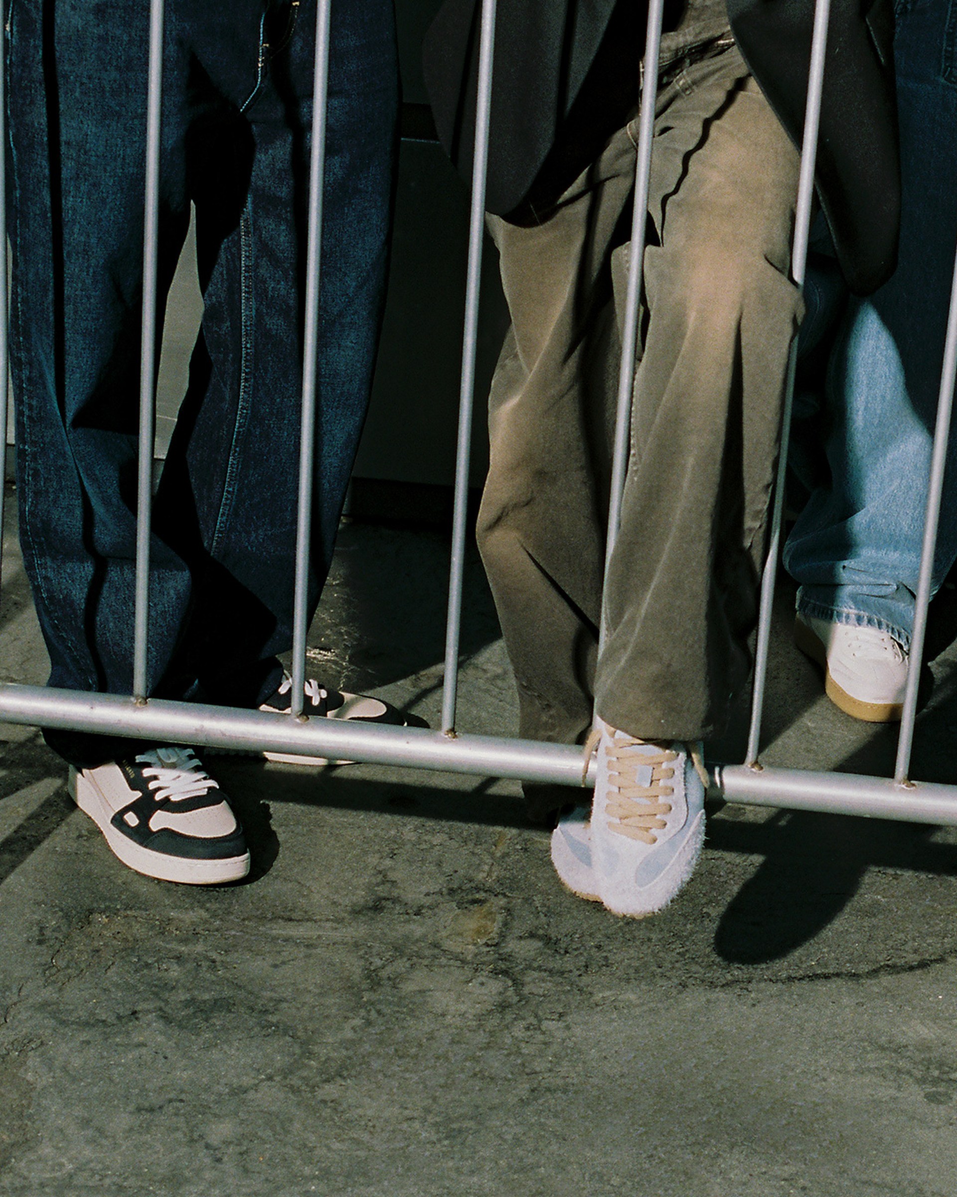 People standing behind a metal barrier, showing only their legs in jeans and sneakers, on a concrete surface.