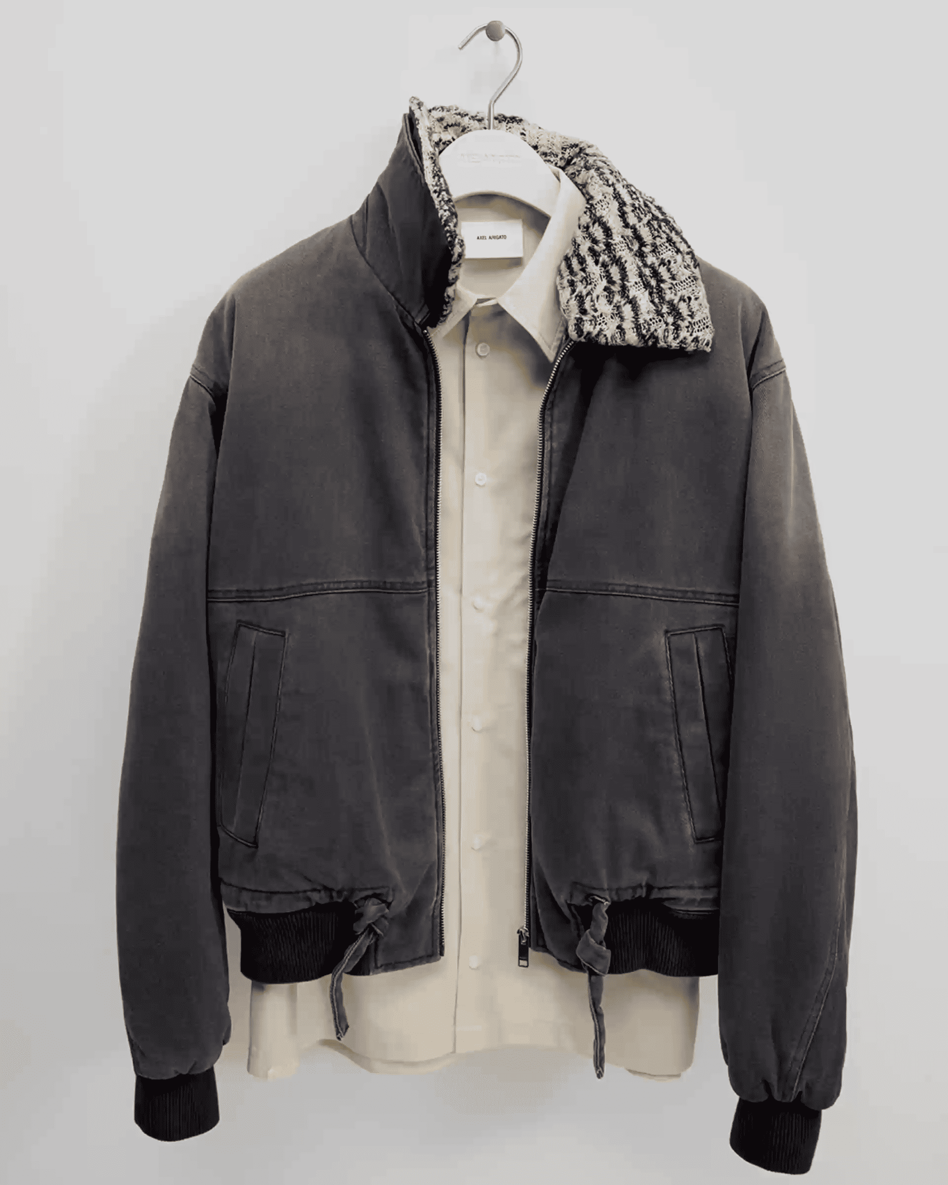 Dark bomber jacket with a patterned collar hung over a beige button-up shirt on a hanger against a light gray wall.