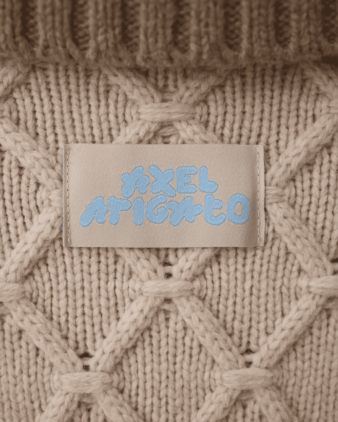 Close-up of a beige knitted sweater with a label displaying the brand name in blue text. The knit has a diamond pattern.