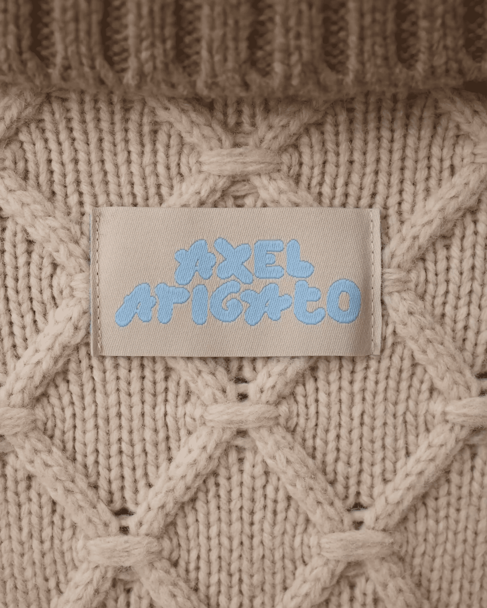 Close-up of a beige knitted sweater with a label displaying the brand name in blue text. The knit has a diamond pattern.