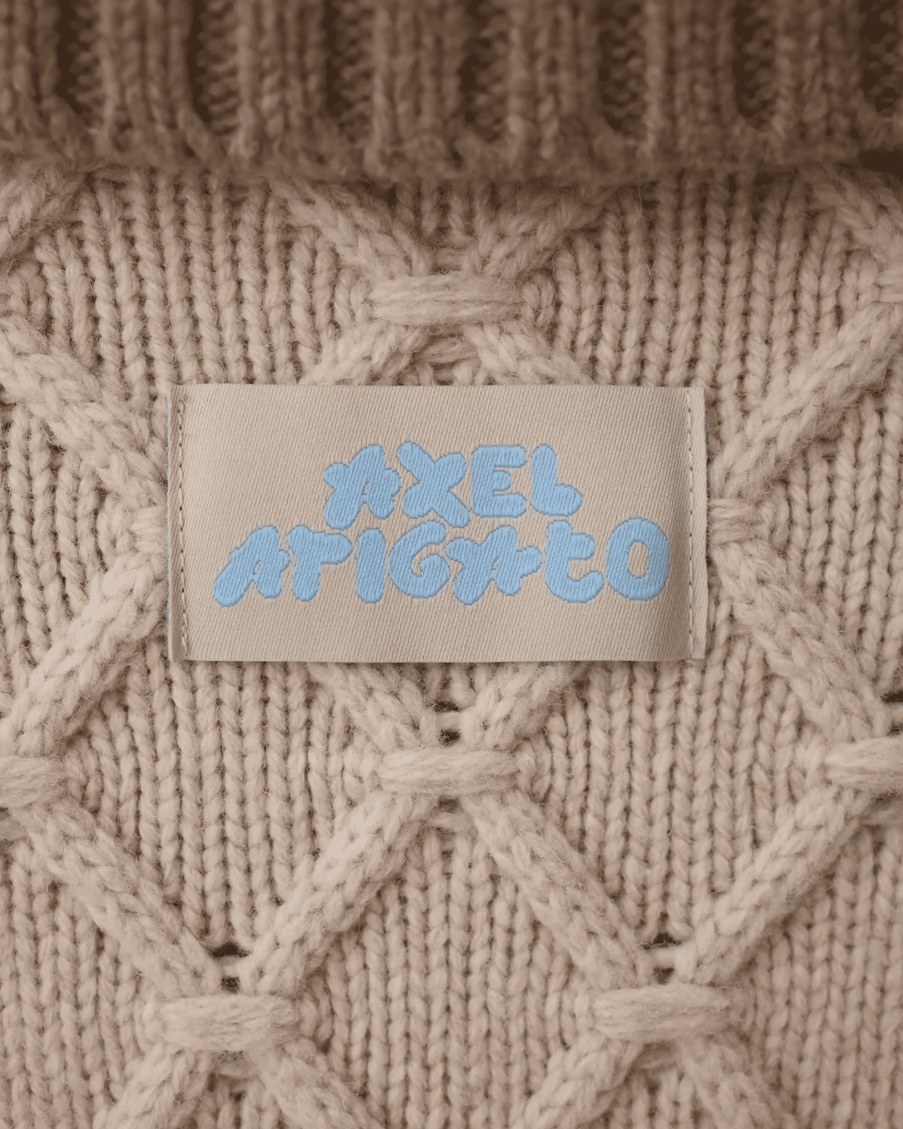 Close-up of a beige knitted sweater with a label displaying the brand name in blue text. The knit has a diamond pattern.
