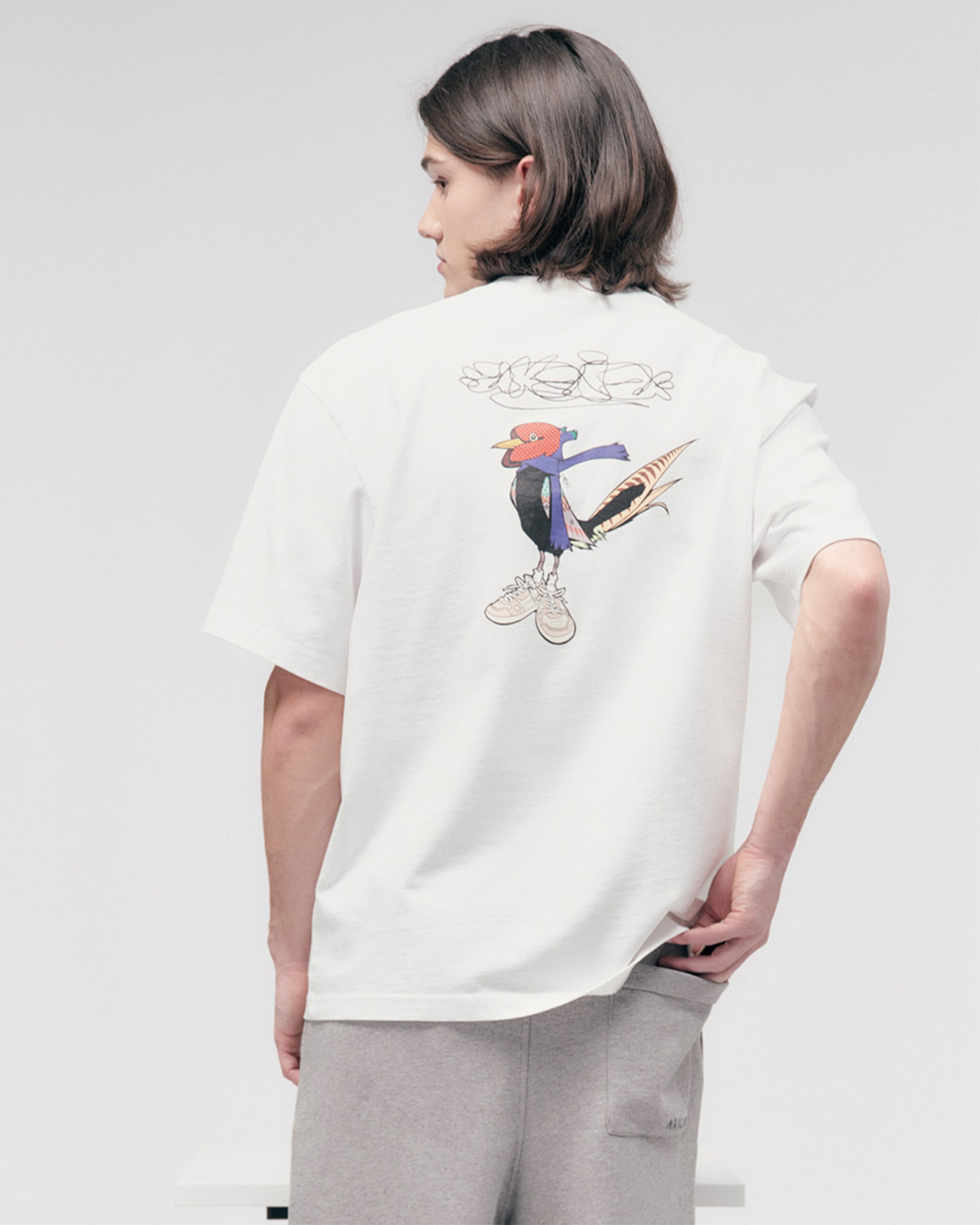 Person with shoulder-length hair wearing a white T-shirt featuring a colorful cartoon design on the back, paired with gray pants.