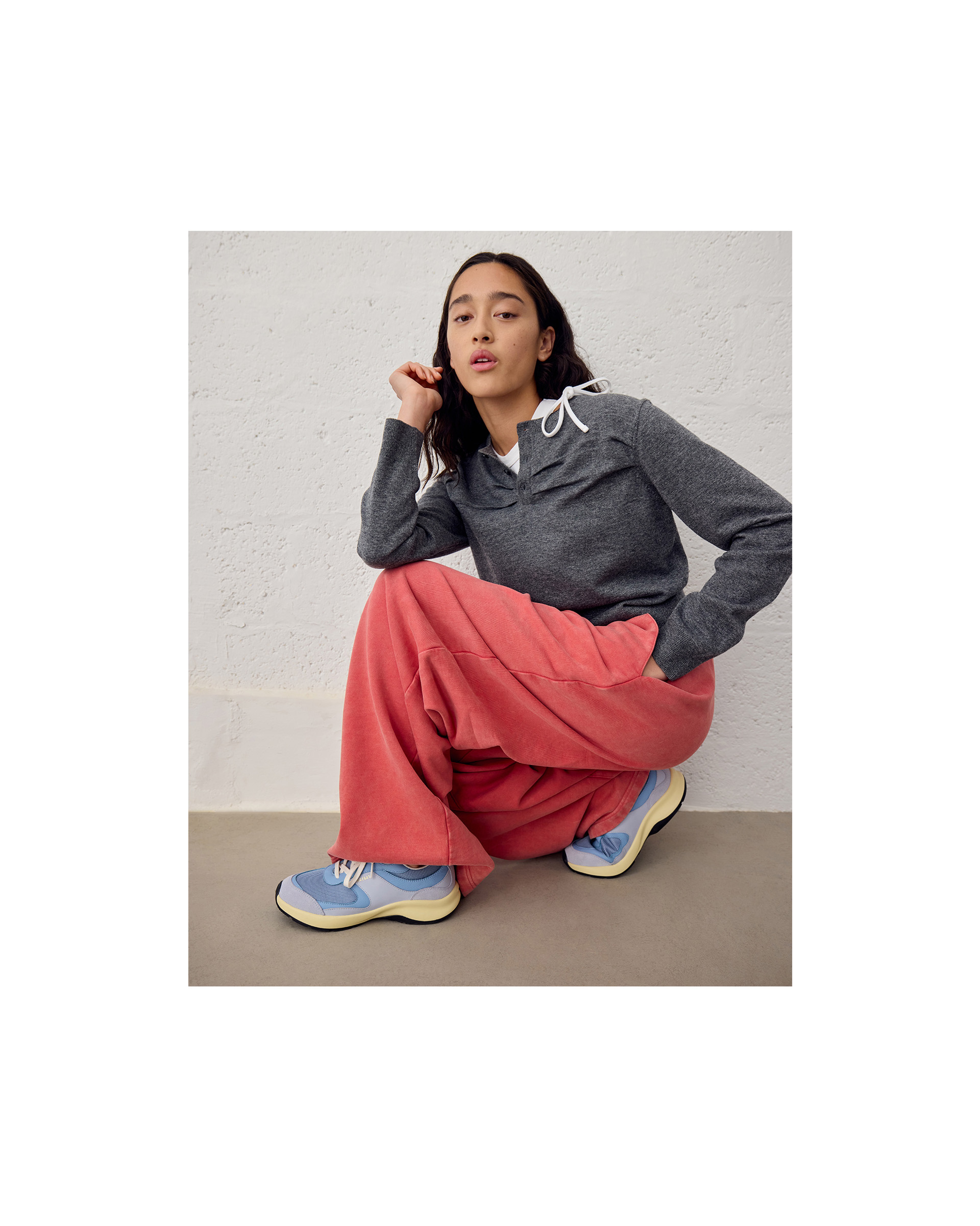 A person squatting against a light wall, wearing a gray hoodie, loose pink pants, and blue sneakers with a neutral expression.