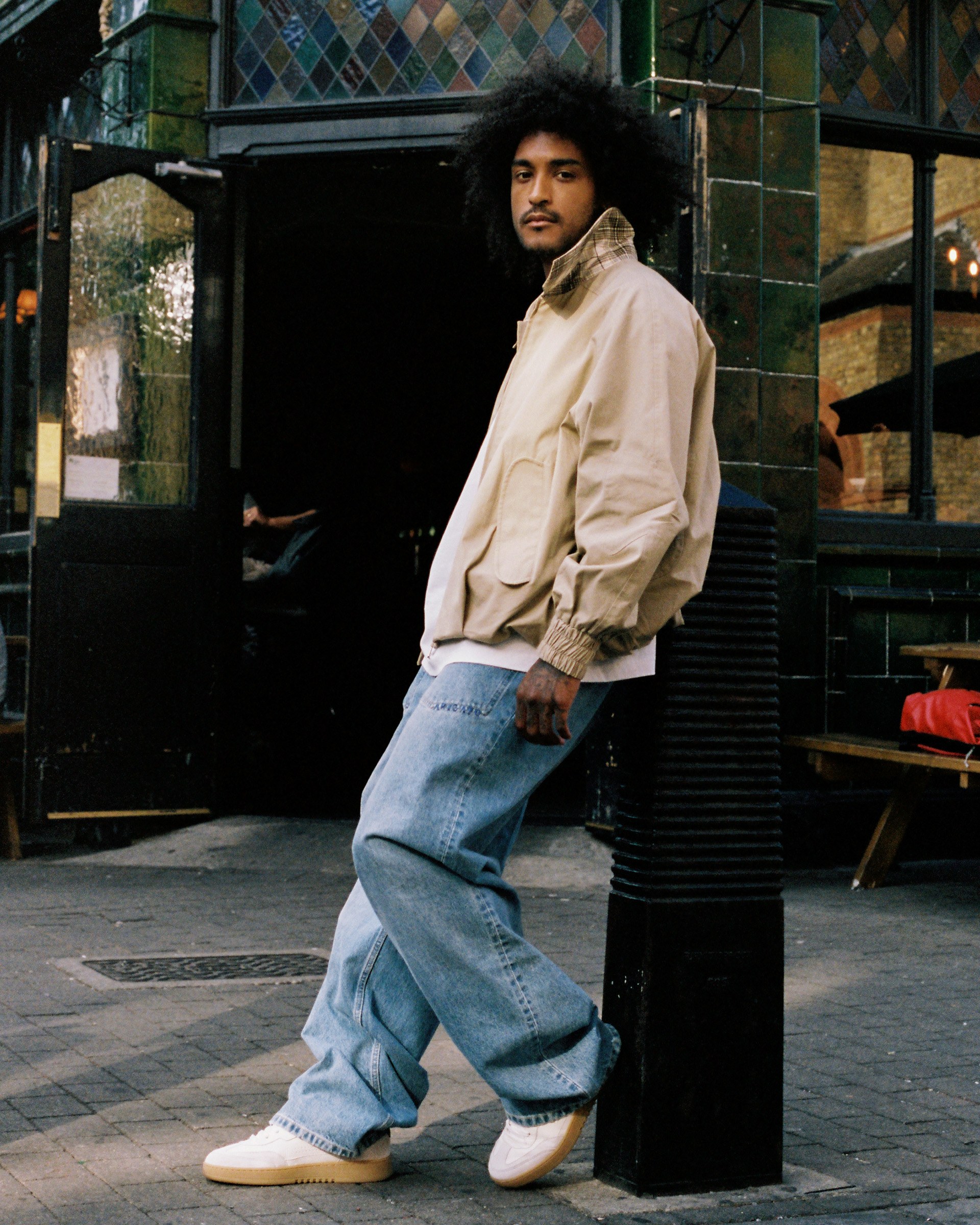 Person with curly hair wearing a beige jacket and blue jeans, standing outside a tiled building with an arched doorway.
