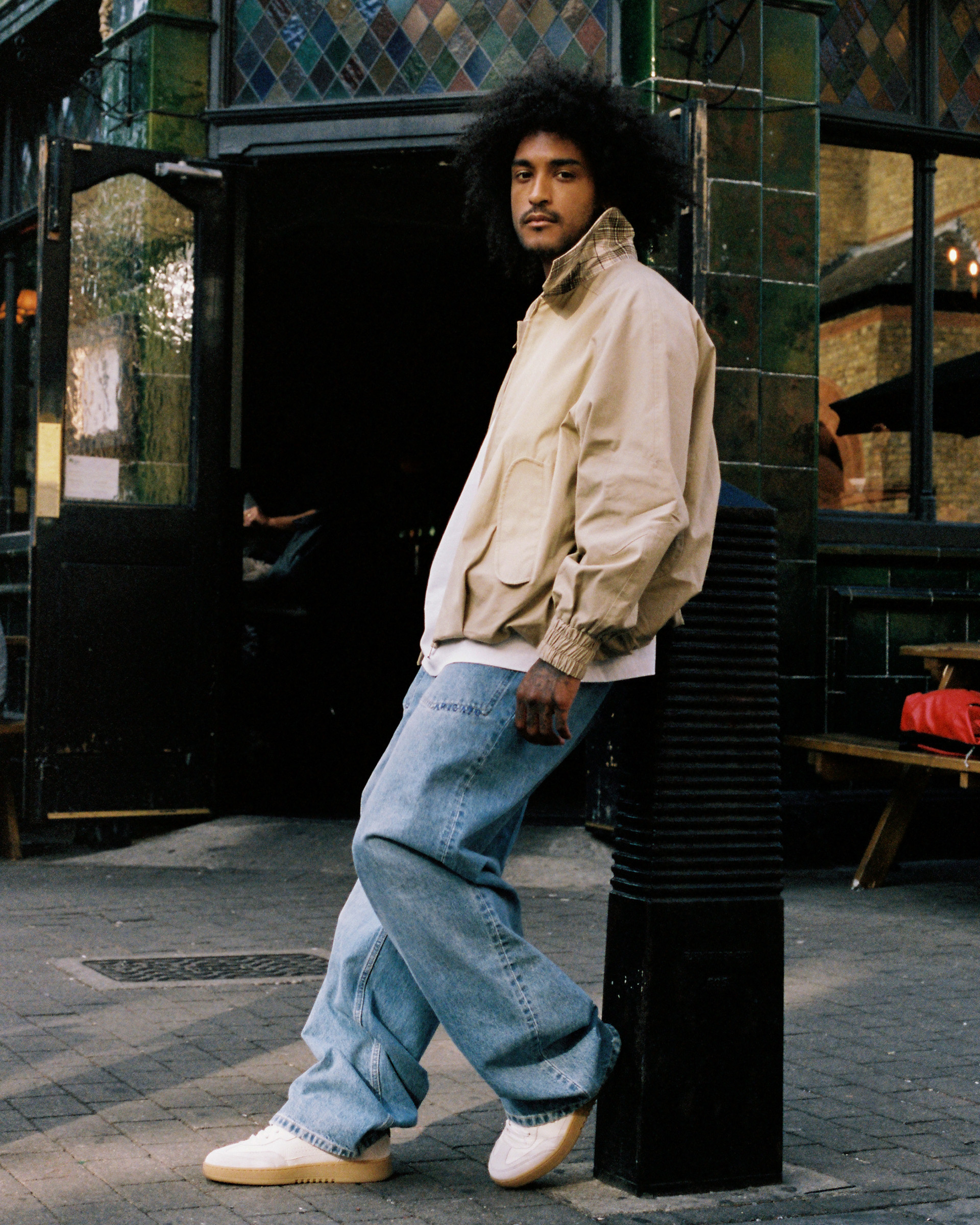 Person with curly hair wearing a beige jacket and blue jeans, standing outside a tiled building with an arched doorway.