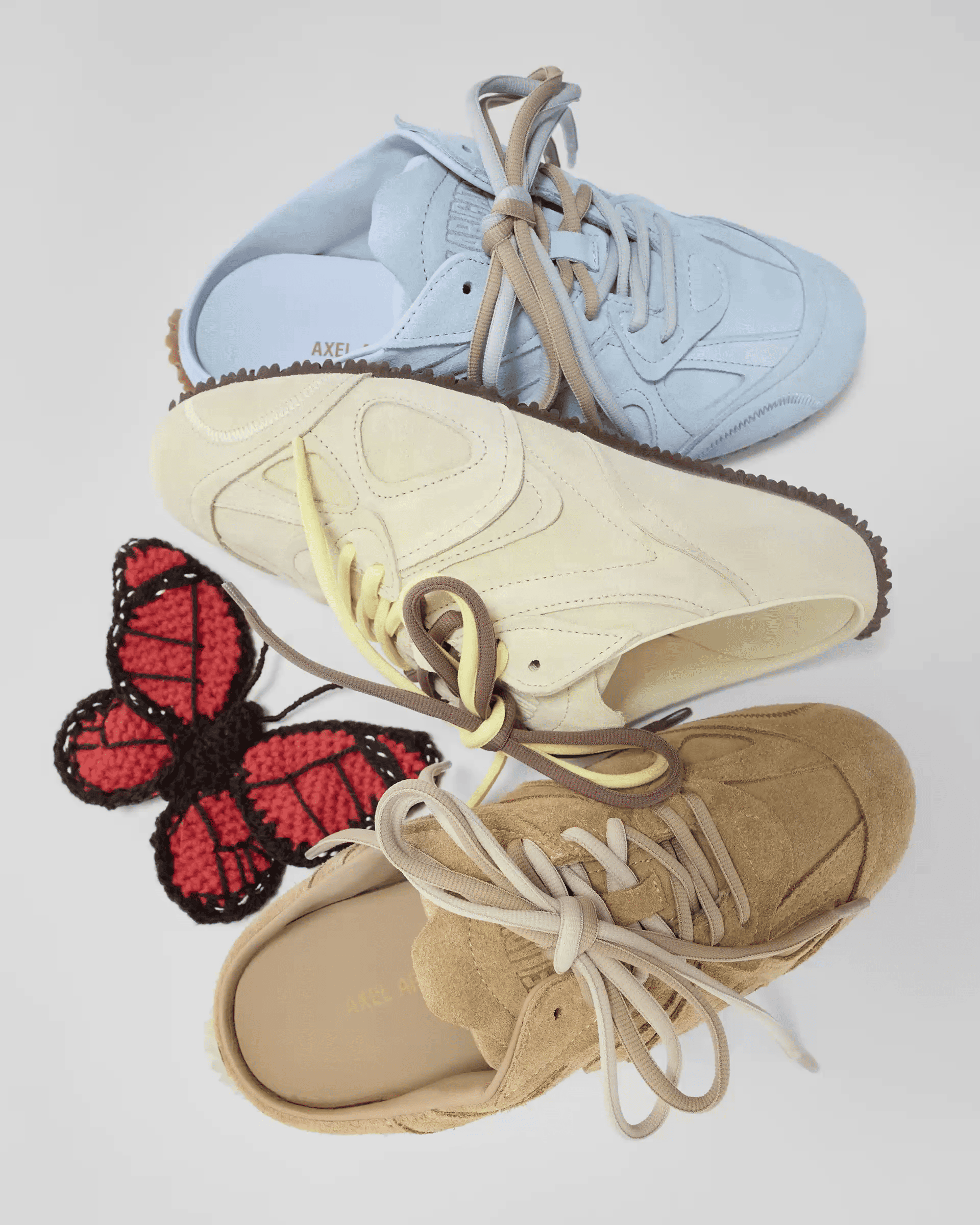 Three sneakers in blue, cream, and tan are displayed with a crocheted red butterfly with black edges on a white background.