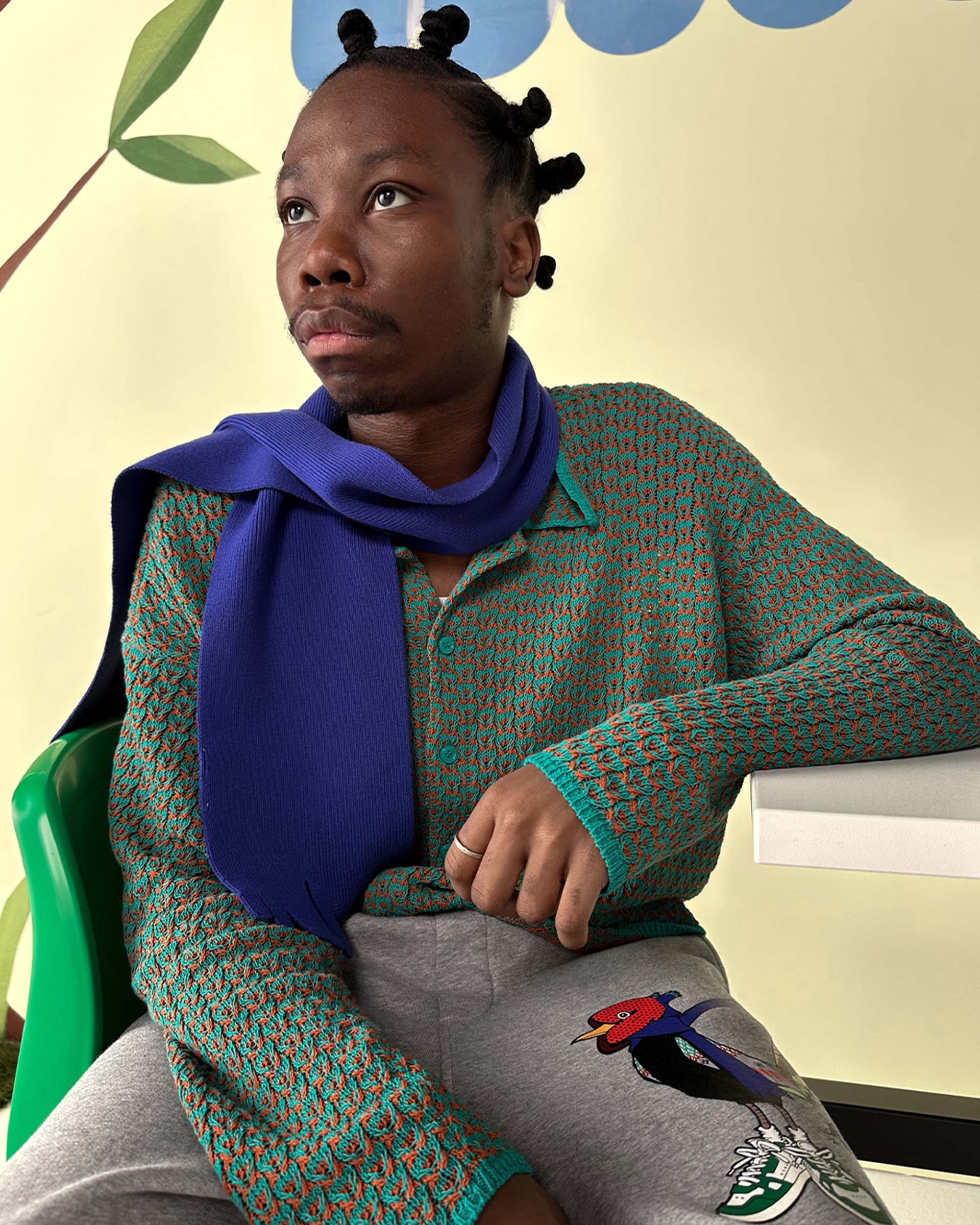 Person with bantu knots, wearing a green and orange sweater, purple scarf, and gray pants with a bird design, sitting on a green chair.