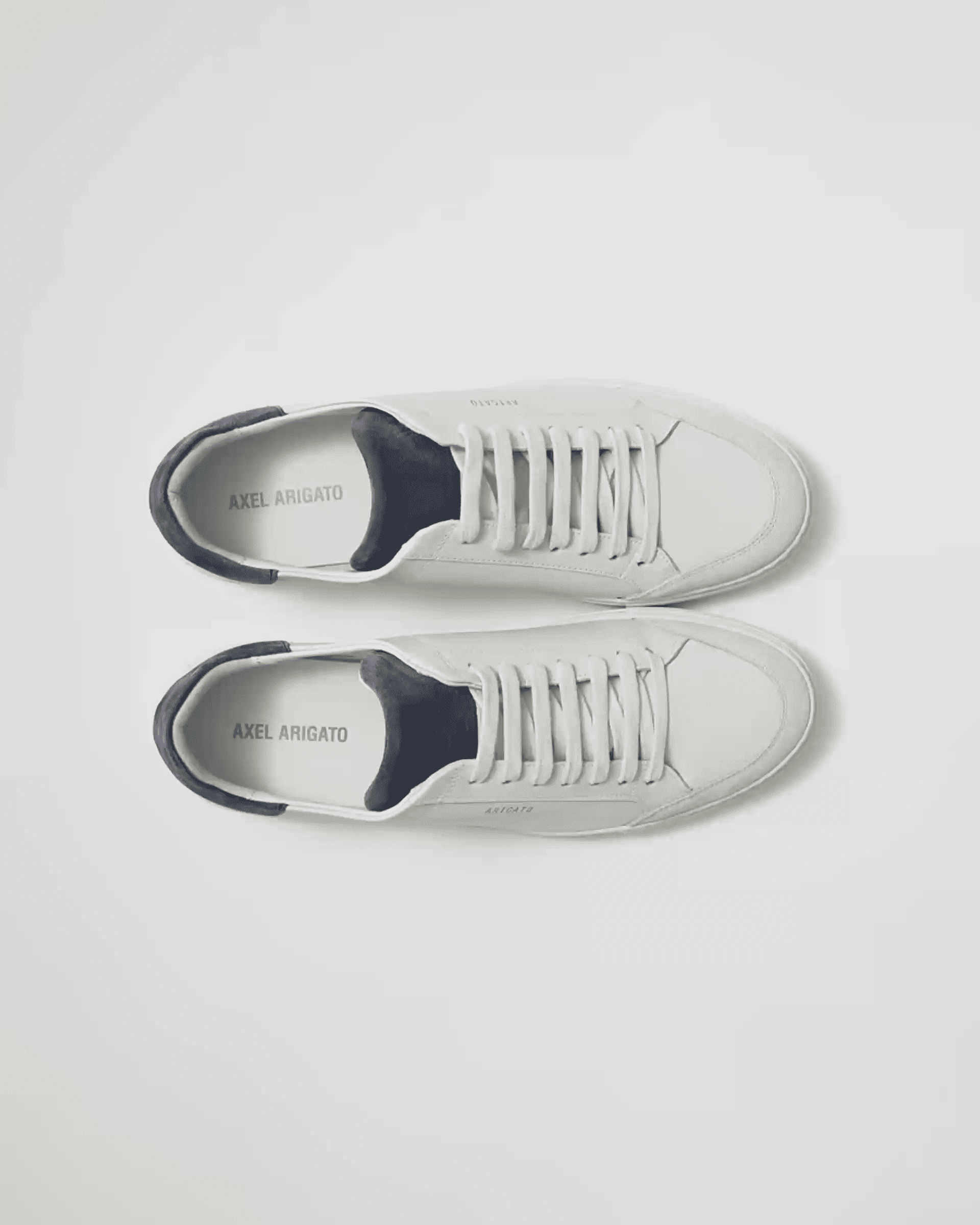 Top view of a pair of white sneakers with black interior and "Axel Arigato" branding on the insoles, laid out side by side.