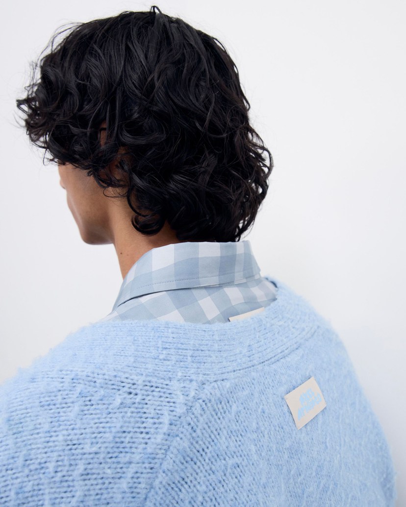 Person with wavy dark hair, wearing a blue checkered shirt under a light blue textured sweater, facing away against a plain background.