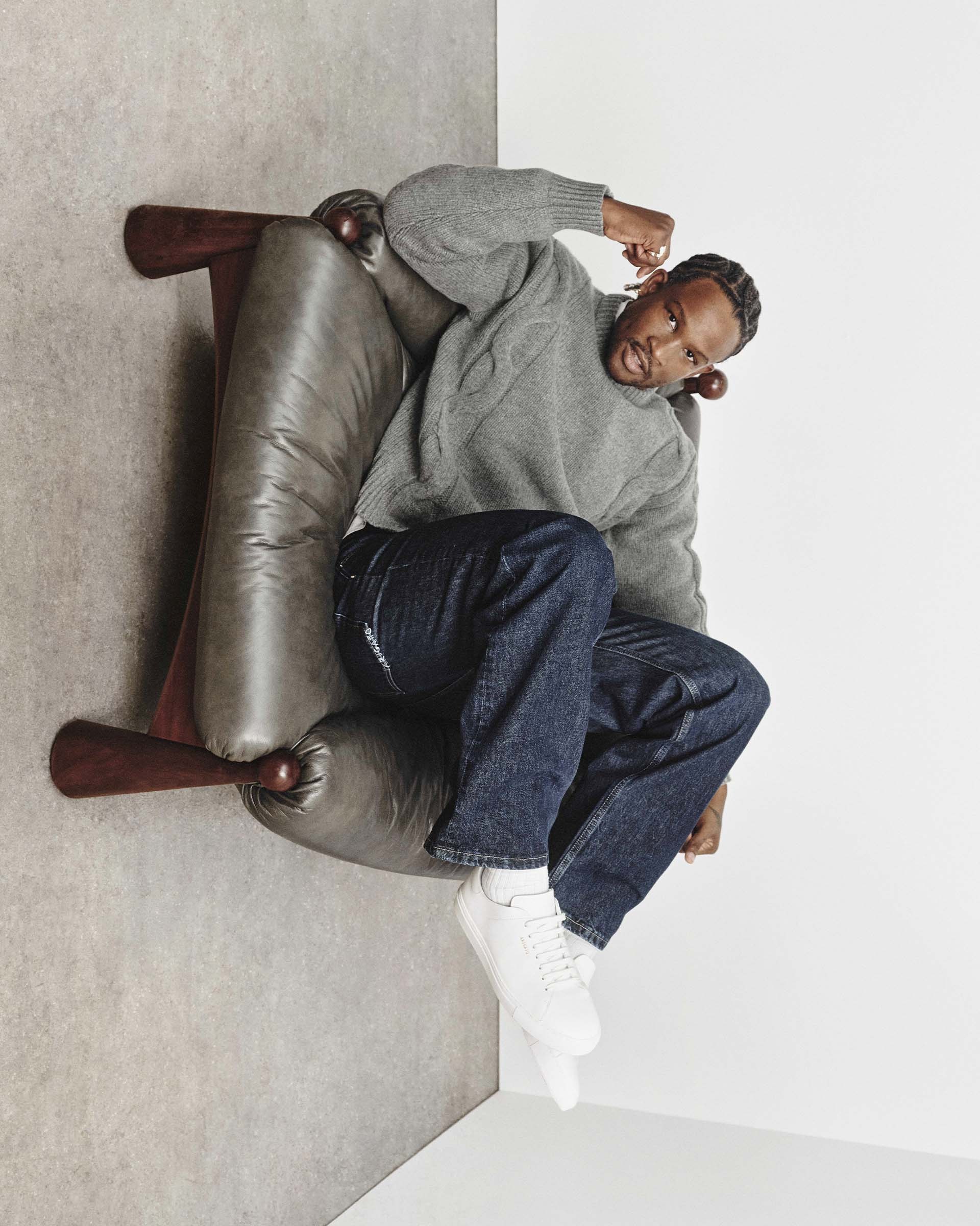 Man in a gray sweater and jeans reclines in an oversized chair, looking relaxed. He wears white sneakers and has braided hair.