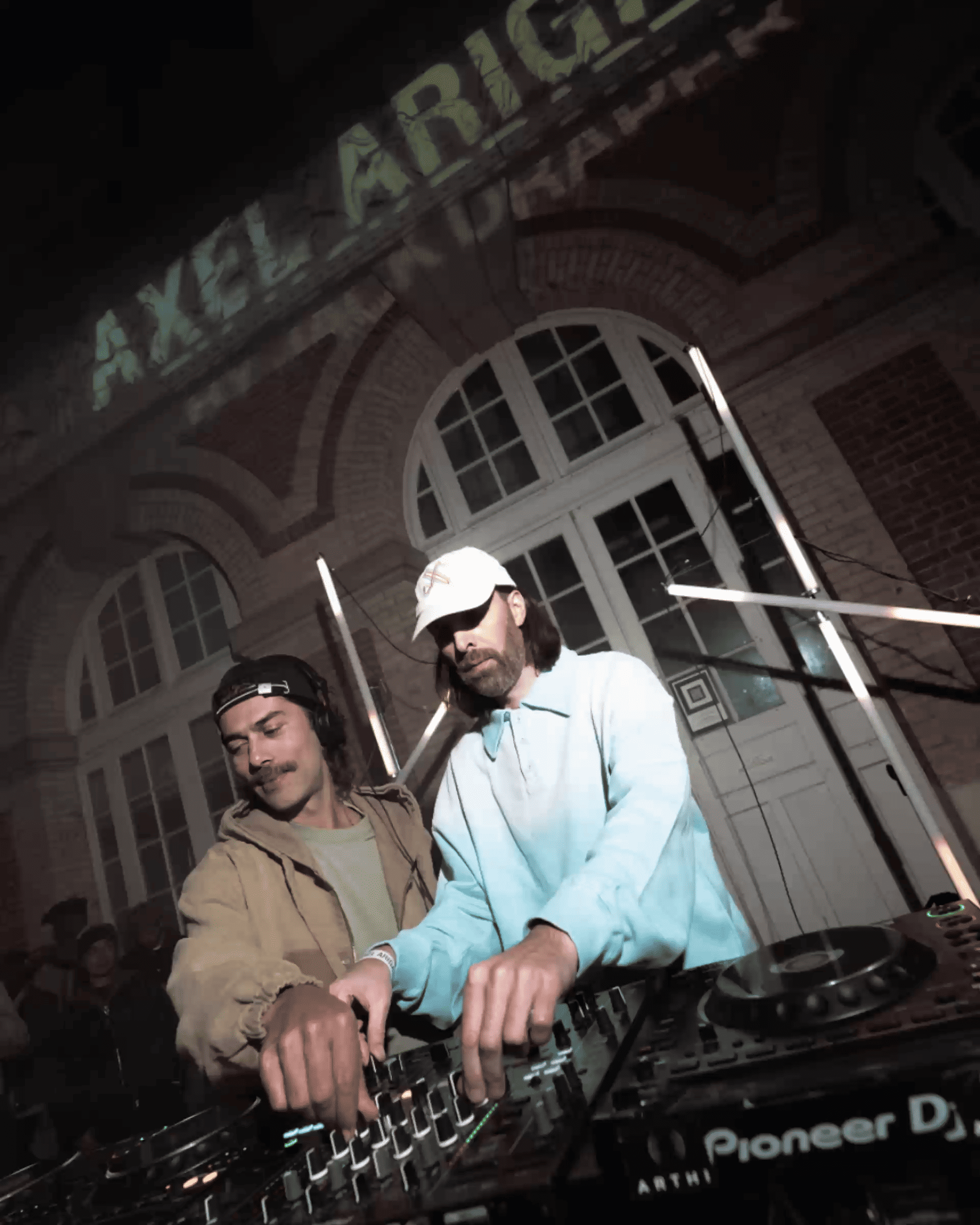 Two DJs mix music together at an event, focused on their equipment. A building with arched windows is lit in the background.
