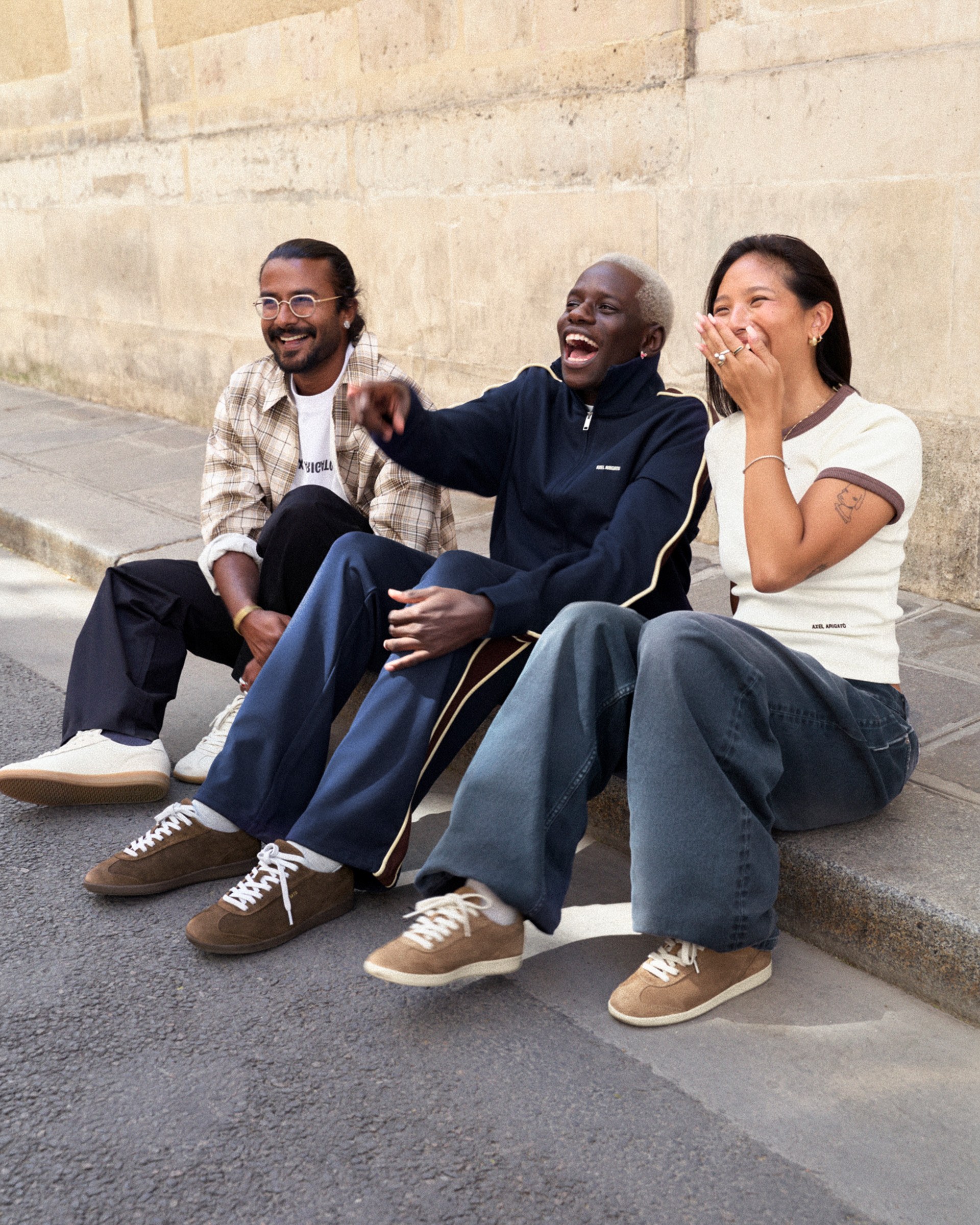 Three people sitting on a sidewalk, laughing and smiling together. They wear casual outfits with sneakers and appear to be enjoying a lively conversation.