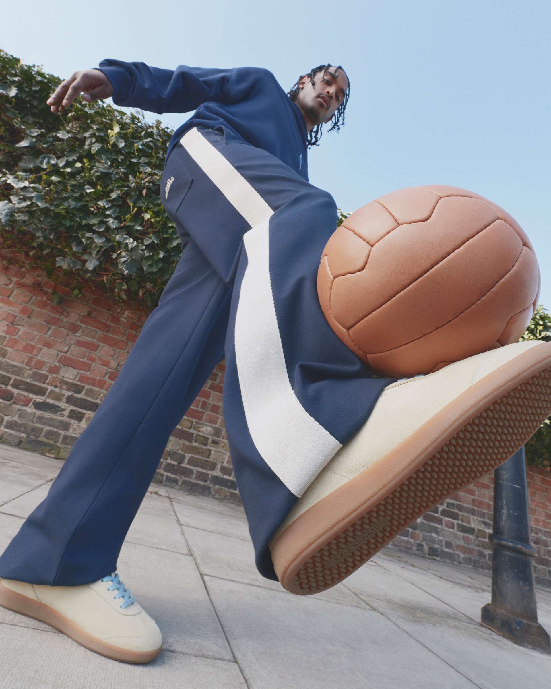 Person in blue tracksuit and beige sneakers skillfully balances a brown soccer ball on their foot, with a brick wall and ivy in the background.